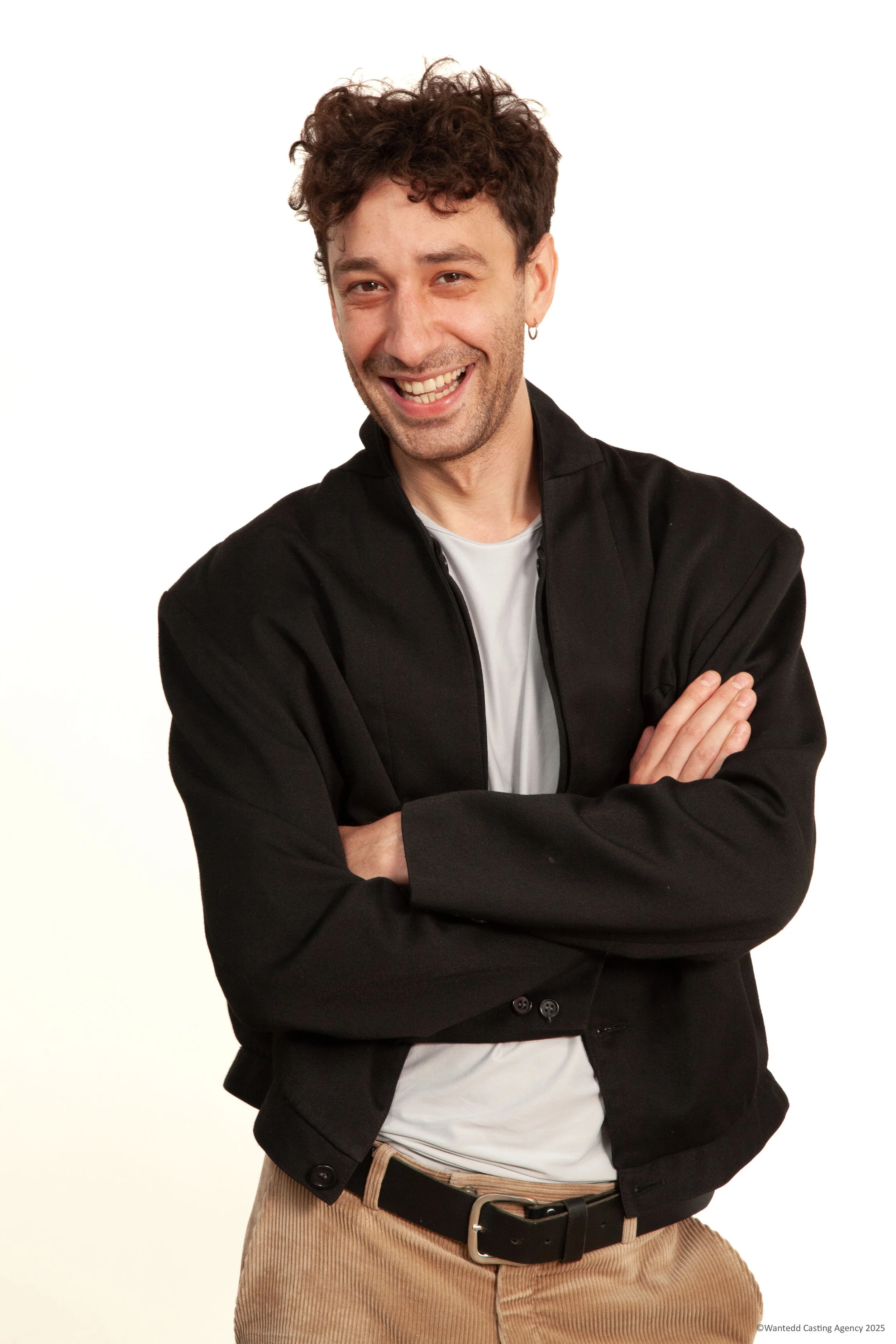 Un homme souriant, aux cheveux bouclés, portant une veste noire et un t-shirt blanc, croise les bras, sur fond blanc.