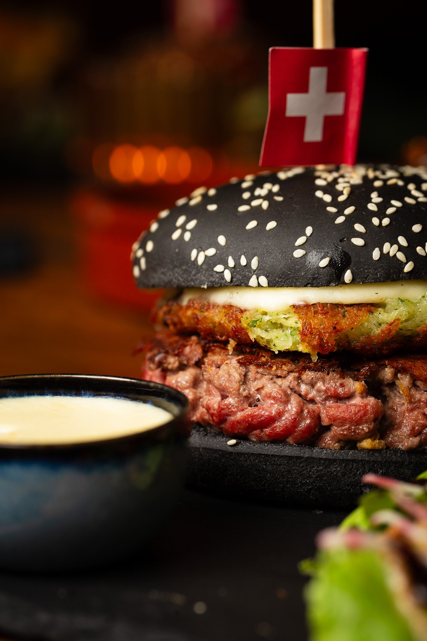 Burger gourmet avec pain noir au sésame, steak, fromage et une galette de pommes de terre, avec un petit drapeau suisse sur le dessus. Une sauce dans un bol à côté.