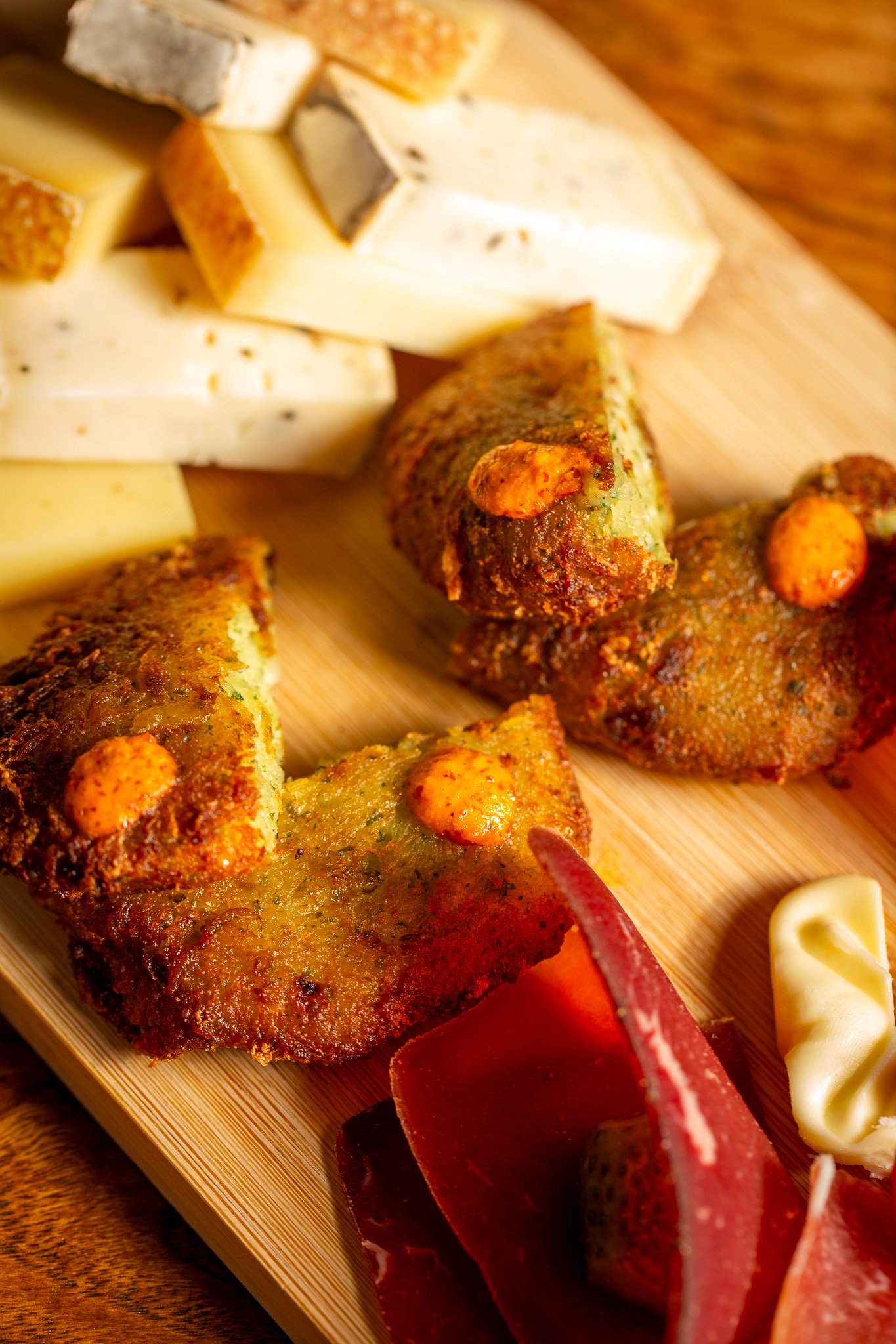 Un plateau de charcuterie et fromage, avec des tranches de fromage variées, des beignets avec sauce épicée, et des morceaux de viande séchée, disposés sur une planche en bois.