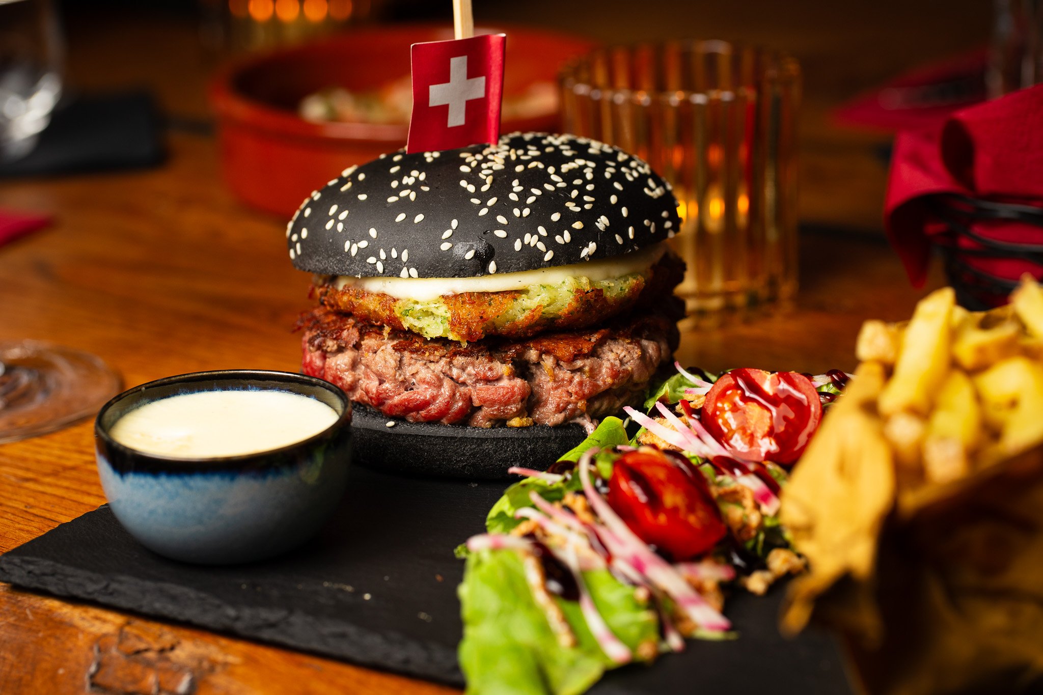Burger à pain noir avec graines de sésame, viande, salade verte, tomates cerises, frites et sauce blanche en accompagnement, décoré d'un drapeau suisse.