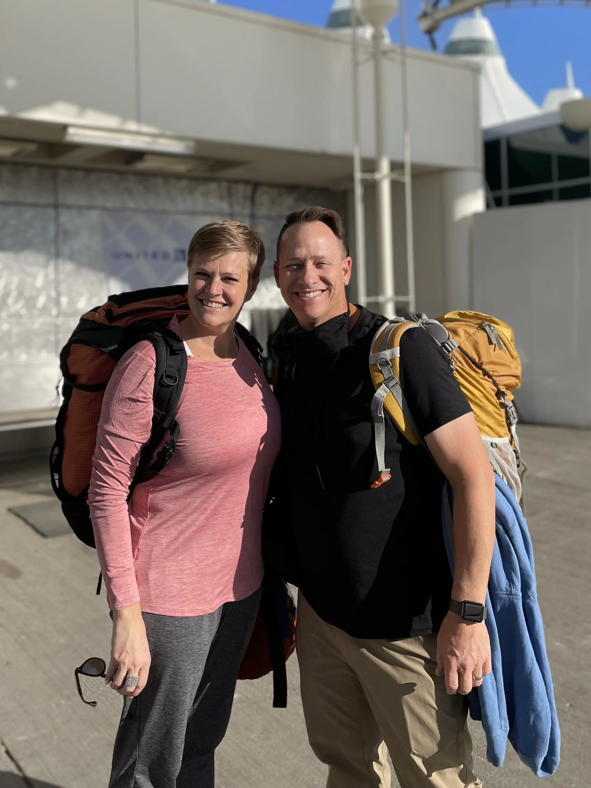 Kristin and Matt smiling, carrying backpacks, standing outside Denver International Airport preparing to leave for Egypt.
