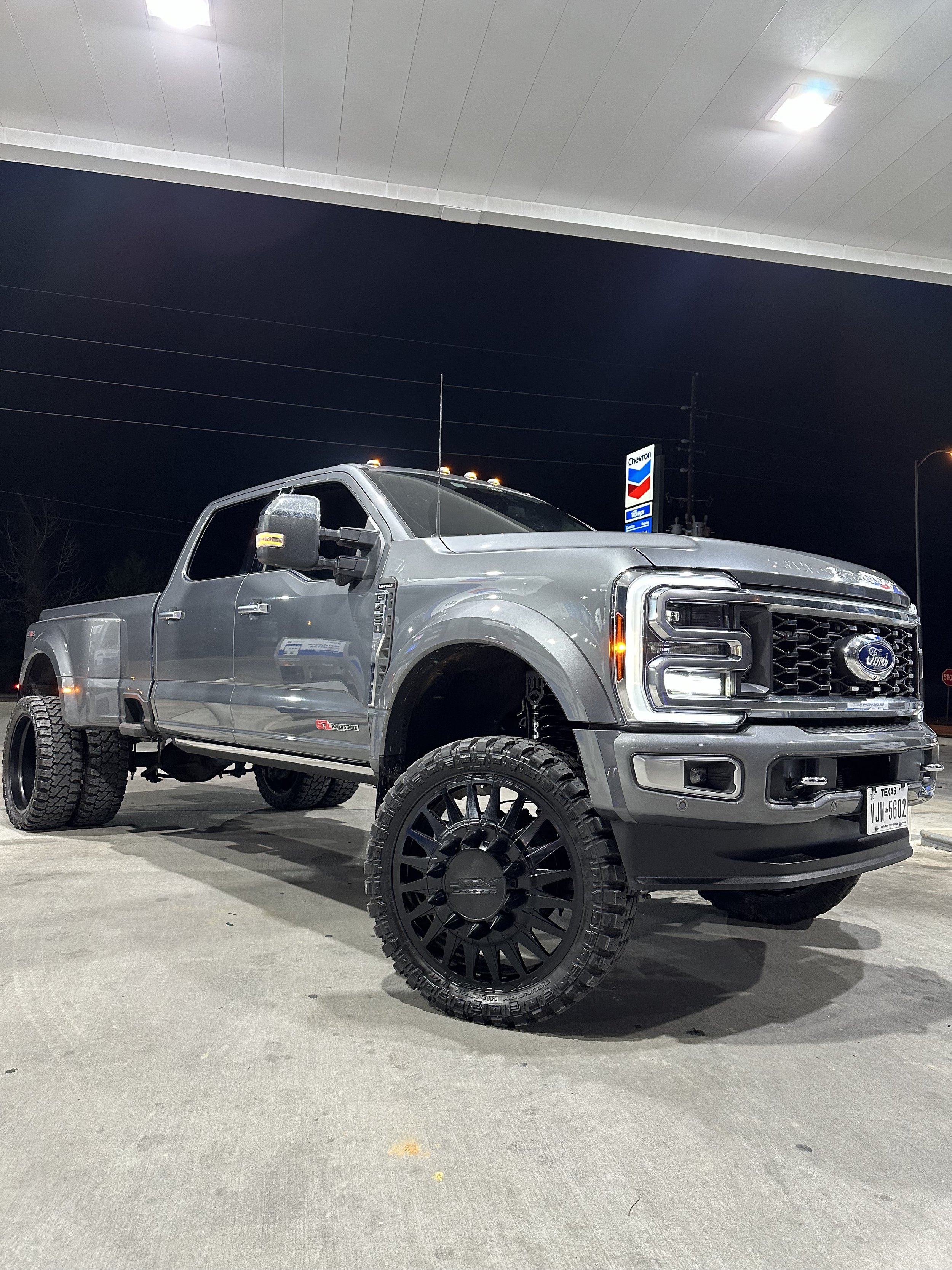 Lifted gray Ford truck with large black wheels at a gas station at night.