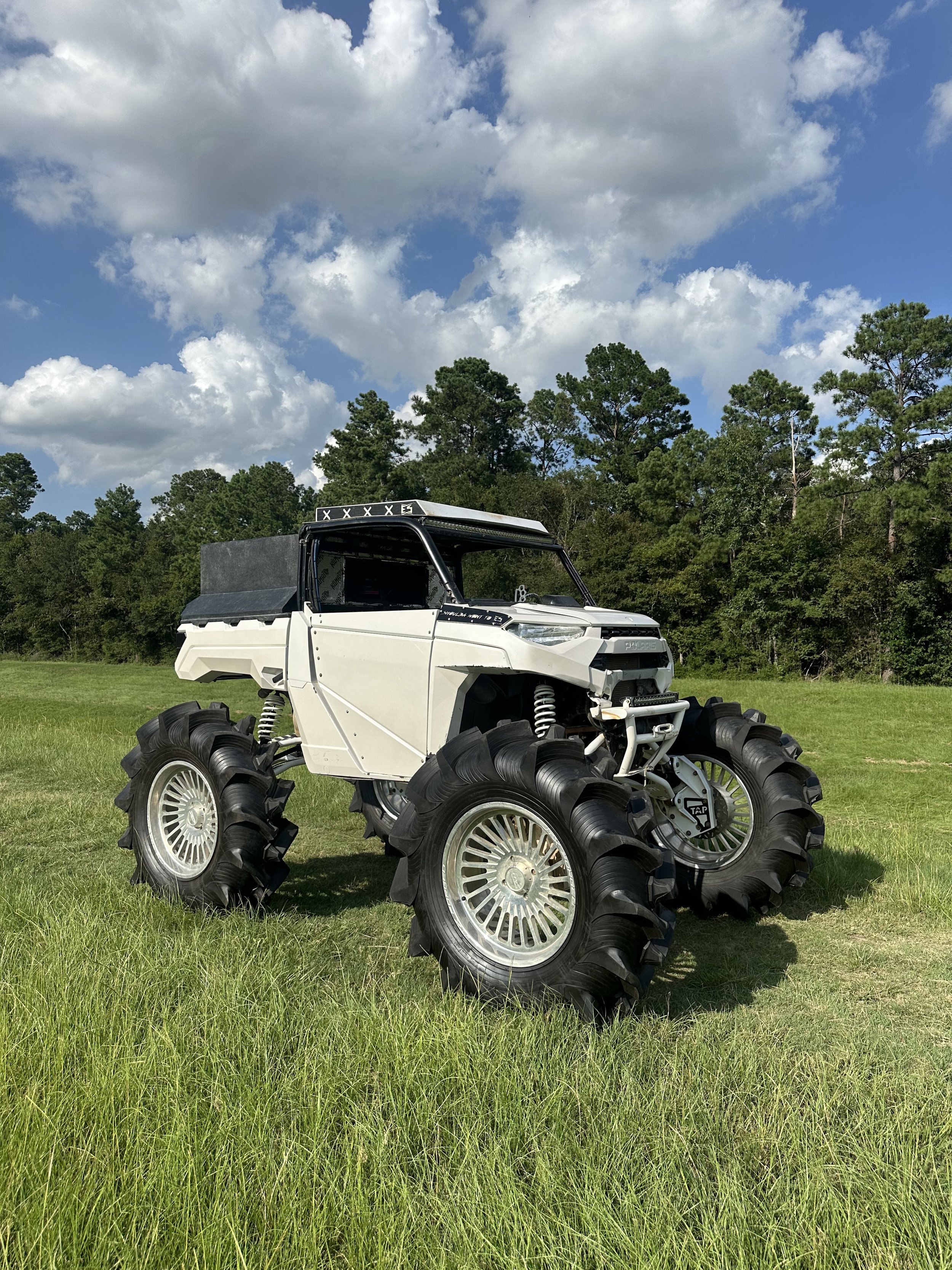 A custom off-road vehicle with oversized tires on a grassy field under a blue sky with clouds.