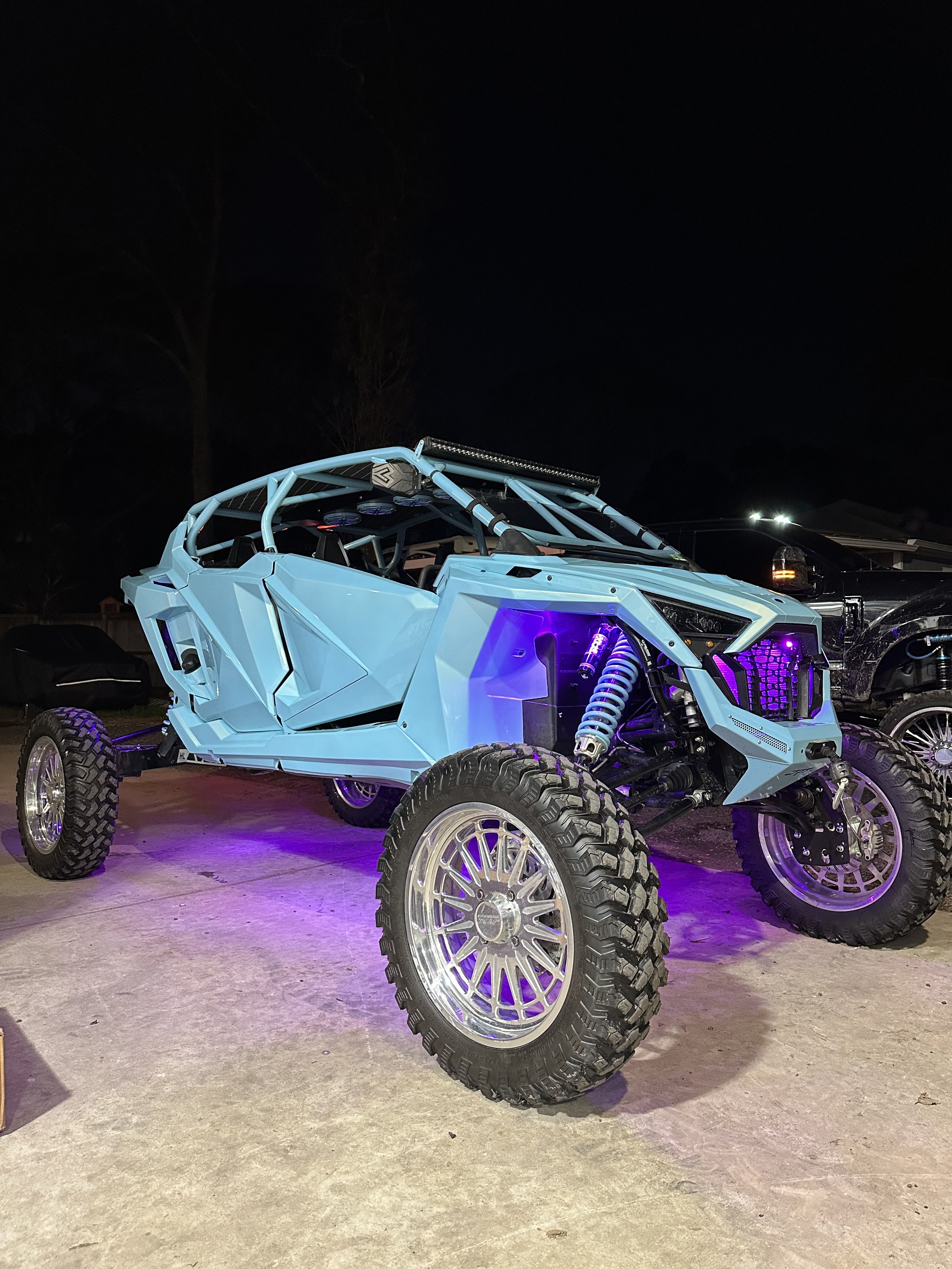 Customized off-road vehicle with large wheels and a light blue cage, illuminated by purple lights, parked on concrete at night.
