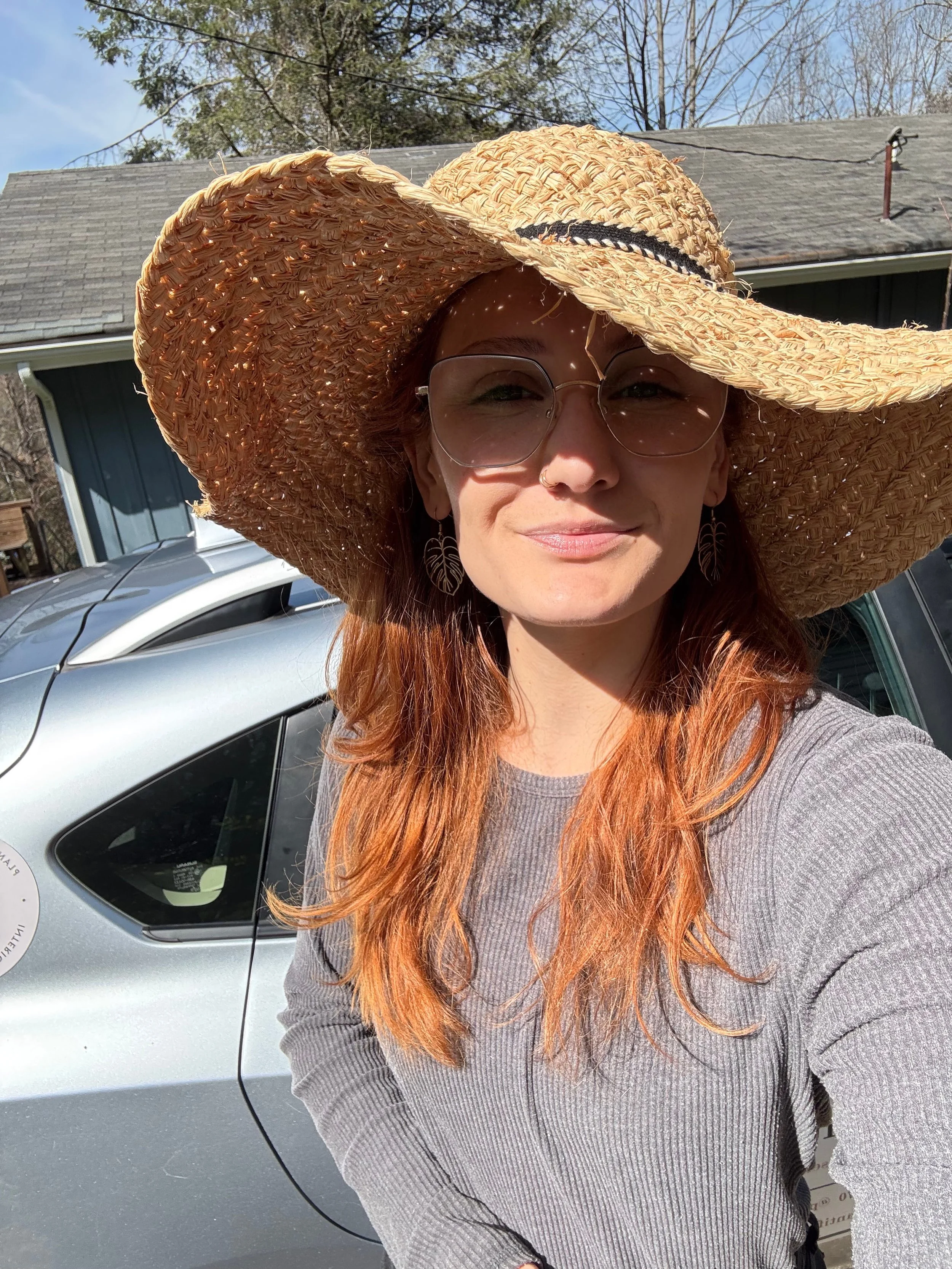 Woman with red hair wearing large straw sun hat, glasses, and leaf-shaped earrings standing outside near a silver car, with a house and trees in the background.