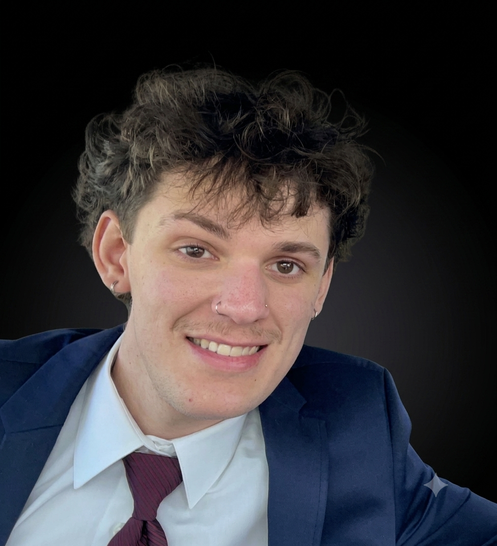 Young person with curly hair, nose rings, wearing a suit and tie, smiling against a black background.