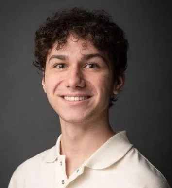 Portrait of a young man with curly brown hair smiling, wearing a white shirt against a dark background.