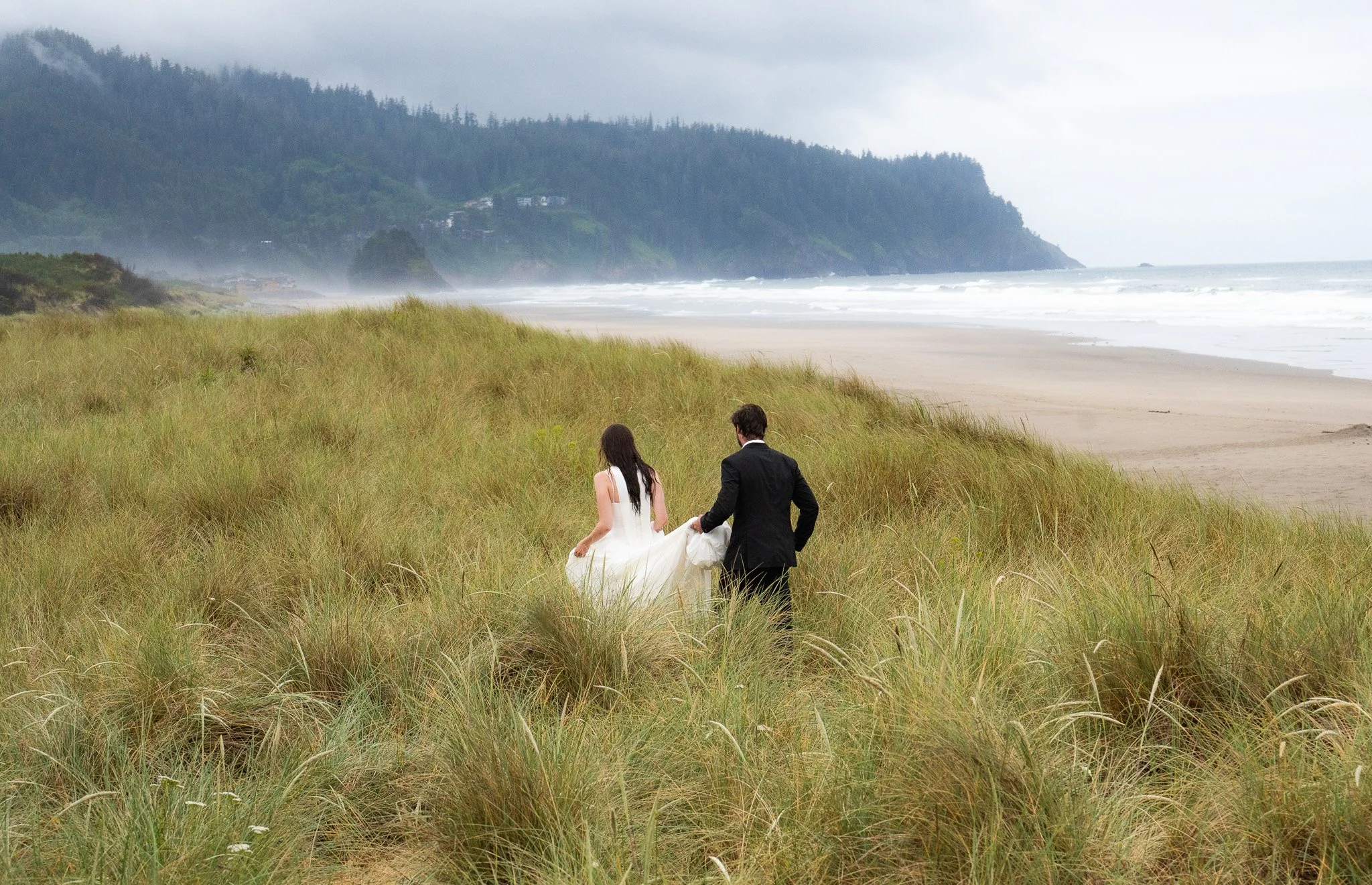 A bride and groom walking on grassy dunes near a beach, with waves and forested hills in the background.