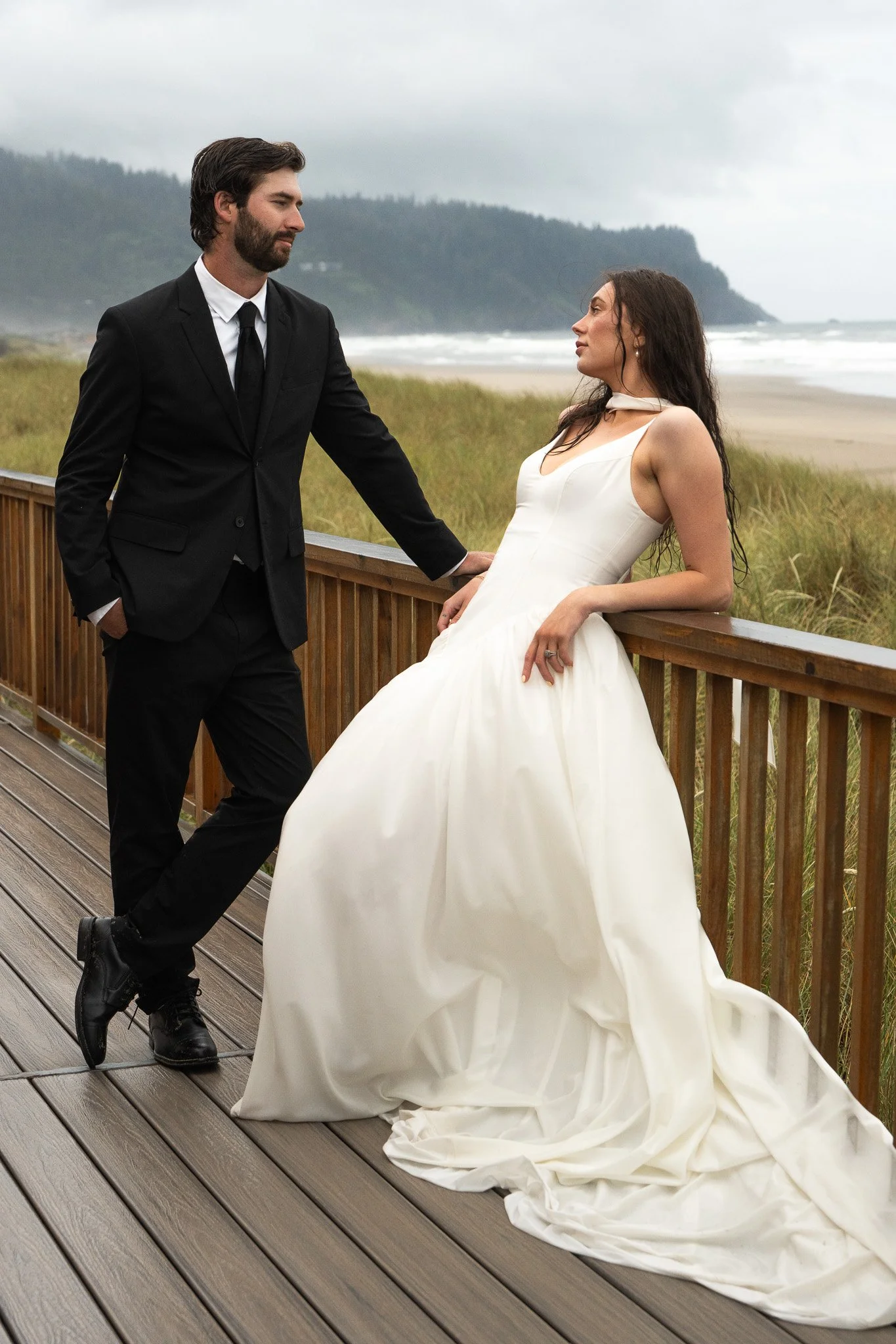 A bride in a white wedding dress and a groom in a black suit and tie pose on a wooden boardwalk near the beach, with grassy dunes and cloudy sky in the background.