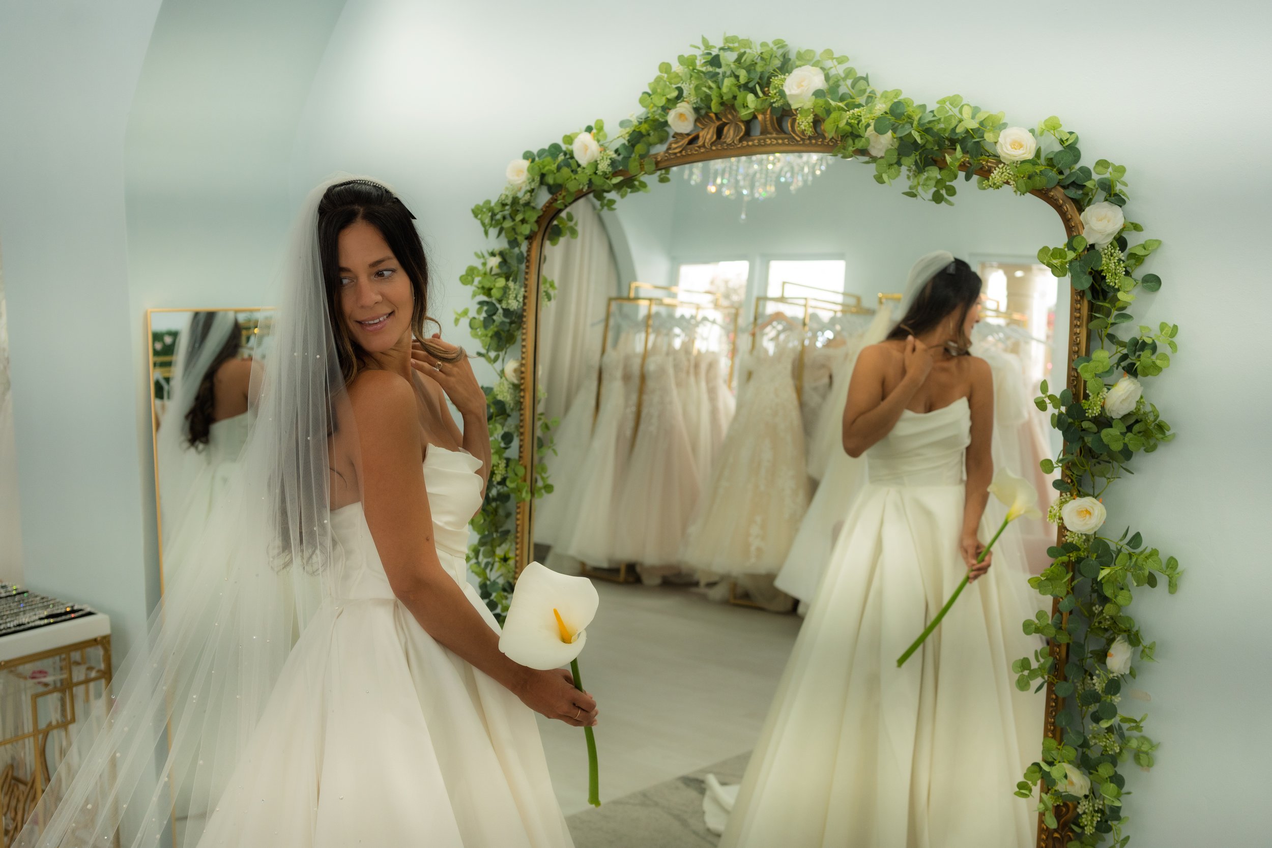 A woman in a wedding dress standing in front of a mirror with a floral garland, holding a calla lily flower, trying on wedding dresses in a bridal boutique.