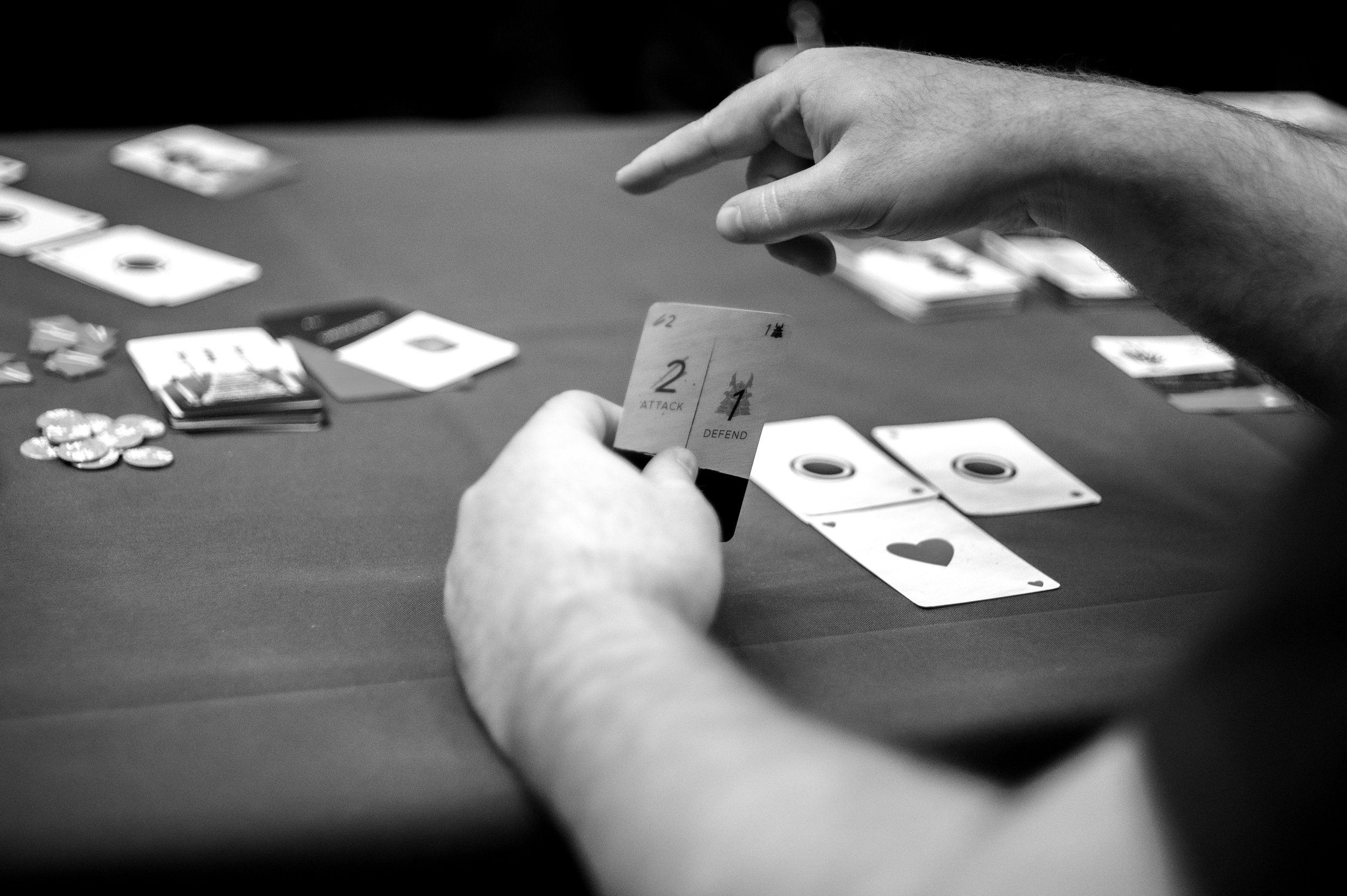 Person playing a card game, holding a card with the words "2 Attack 1 Defend," with poker chips and other cards on a table.