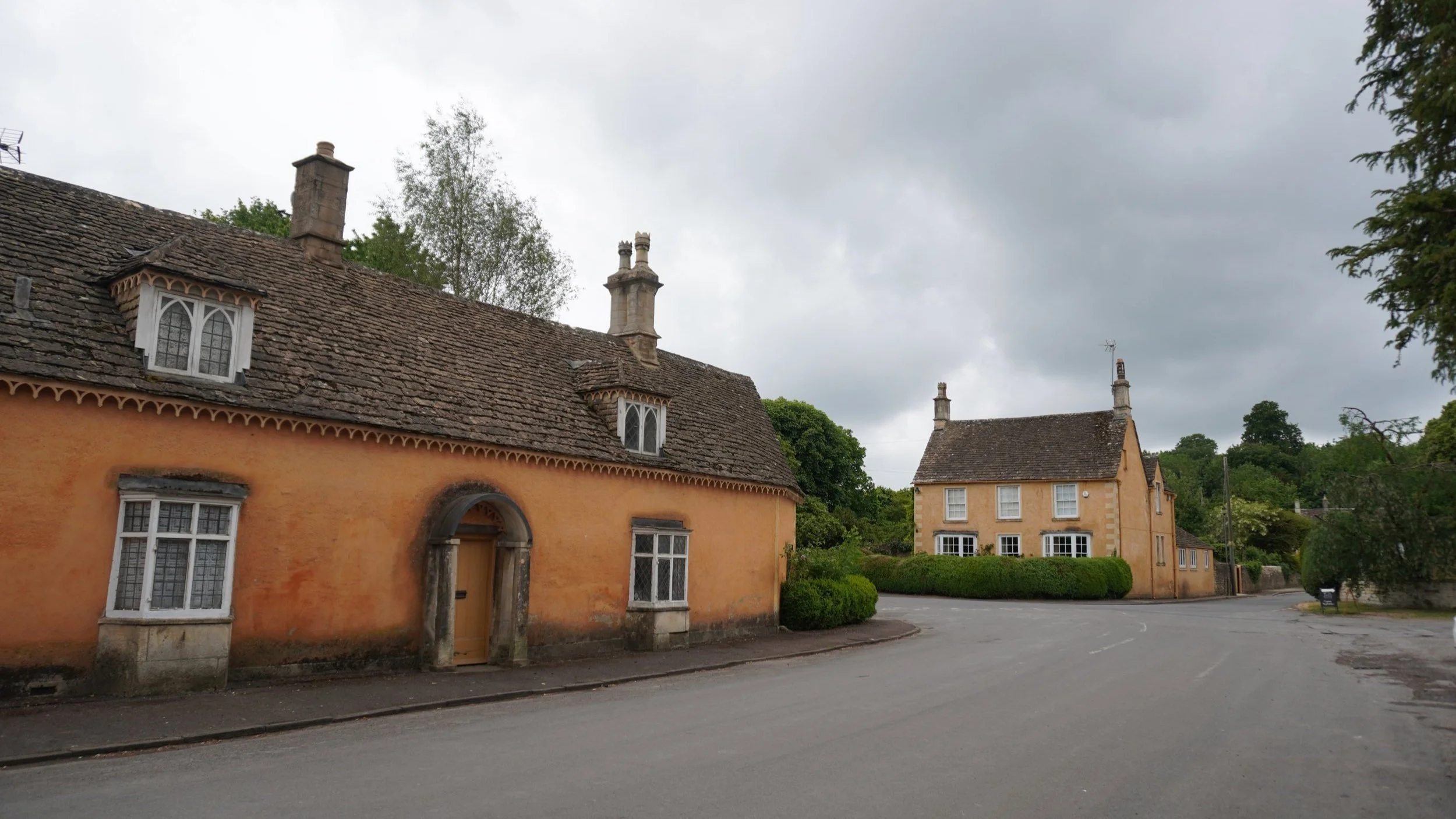 Traditional ochre-washed stone cottages with Cotswold stone tiled roofs and dormer windows along a curving street in Badminton village, Gloucestershire