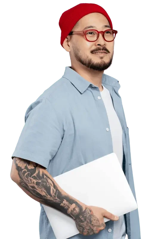 A man with a beard, glasses, red beanie, and light blue shirt holding a white laptop, with tattoos on his arm, smiling slightly.