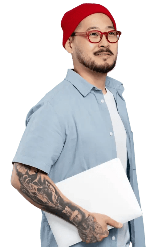 Man with a beard, glasses, red beanie, wearing a light blue button-up shirt, holding a closed laptop, with tattoos on his left arm, standing against a black background.