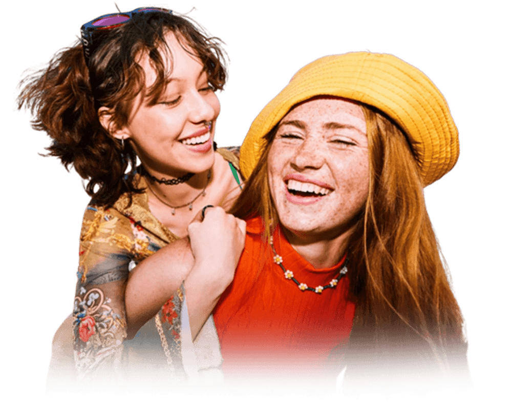 Two women smiling and laughing, one with curly brown hair and sunglasses on her head, the other with long red hair wearing a yellow hat and a red top.