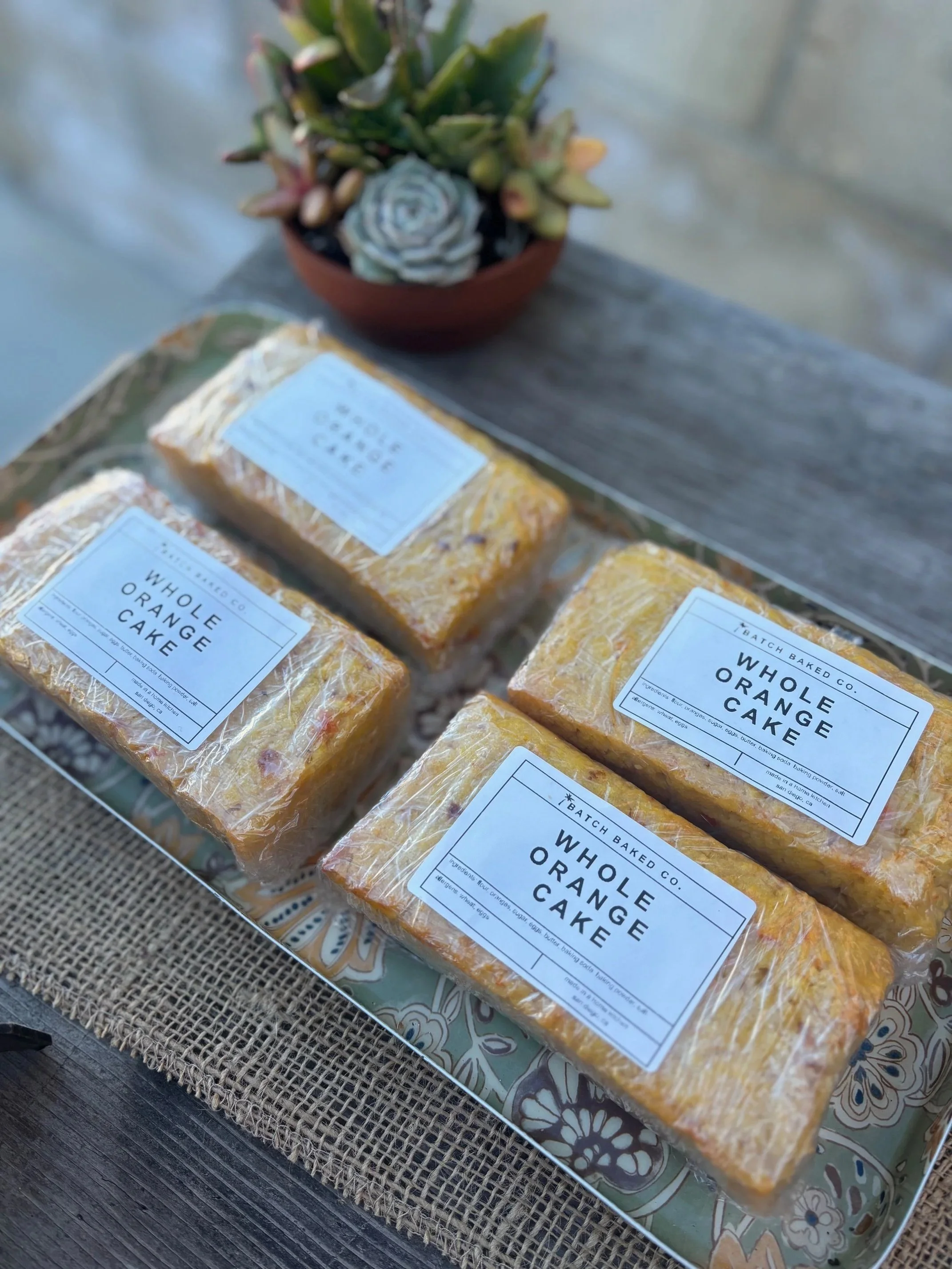 Four loaves of batch baked whole orange cake wrapped in plastic, placed on a decorative tray, with a small potted succulent plant in the background.