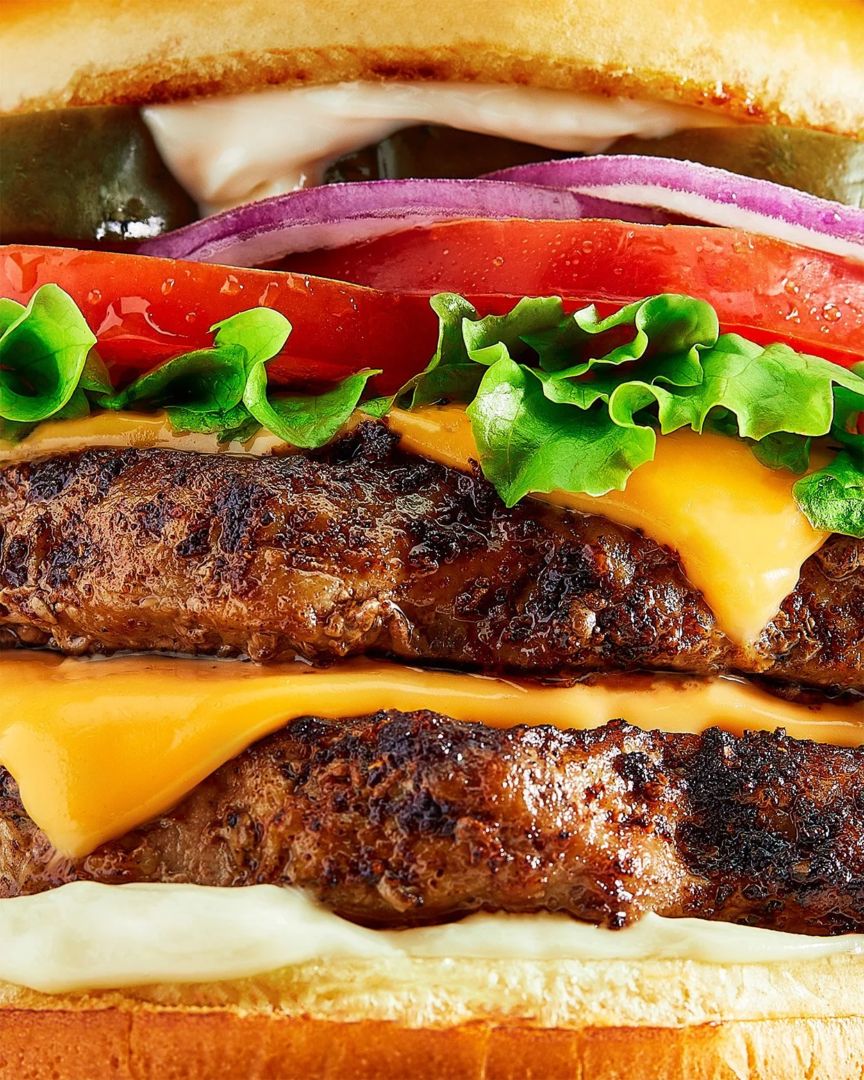 Close-up of a cheeseburger with beef patty, cheddar cheese, lettuce, tomato, onion, pickles, and mayonnaise on a bun.