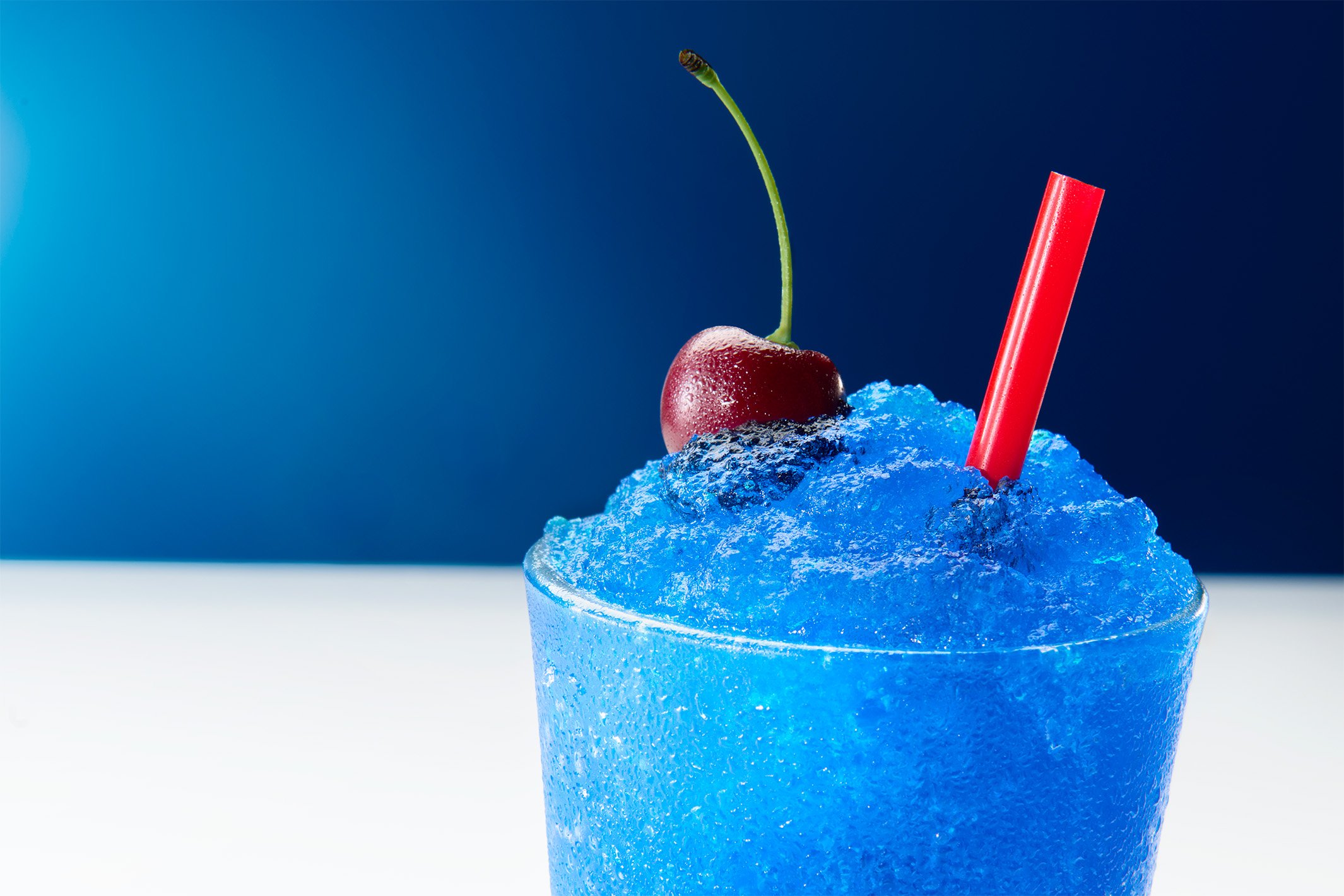 Close-up of a blue icy beverage in a glass with a cherry and a red straw on a white surface against a blue background.