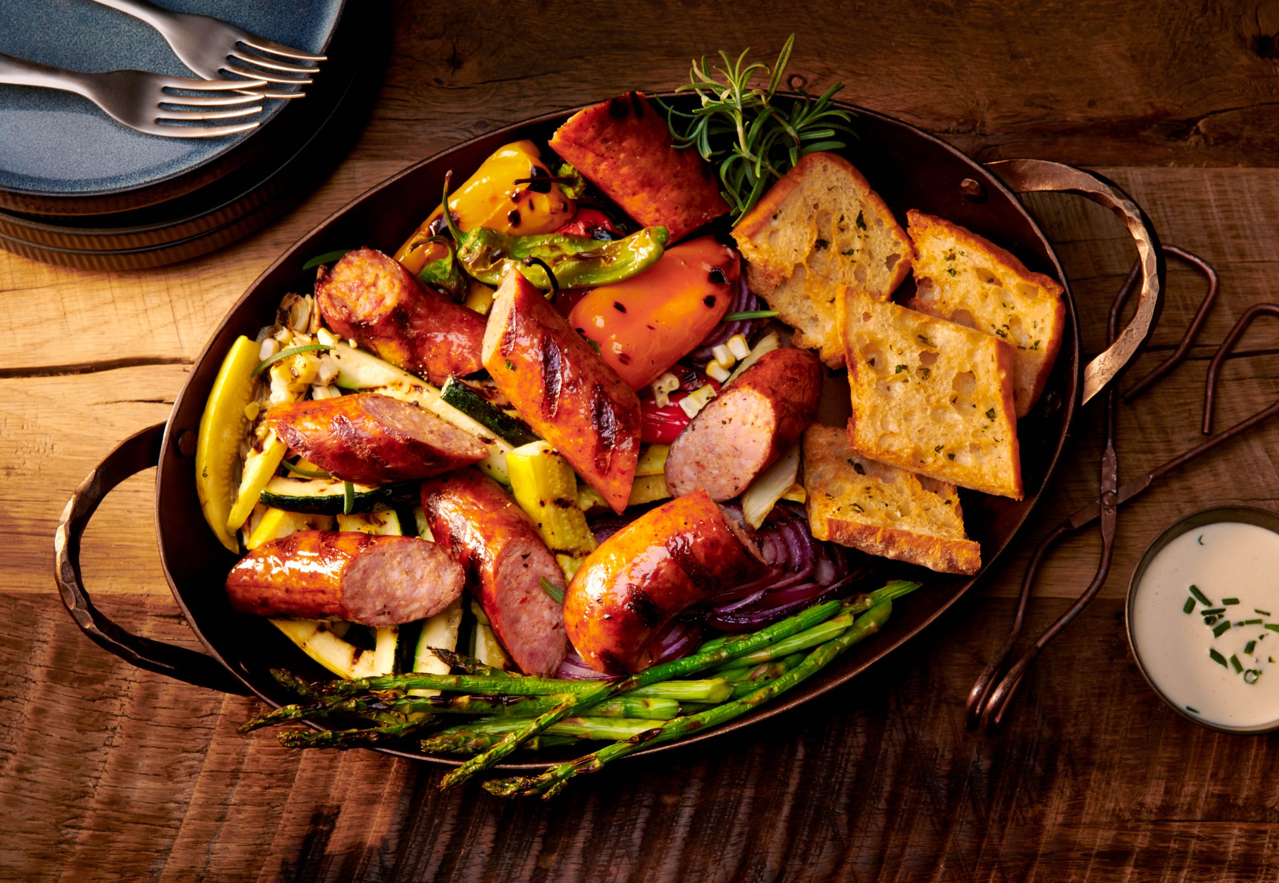 A rustic dinner plate with grilled sausages, roasted vegetables, slices of garlic bread, and garnished with fresh herbs, served with a side of dipping sauce on a wooden table.