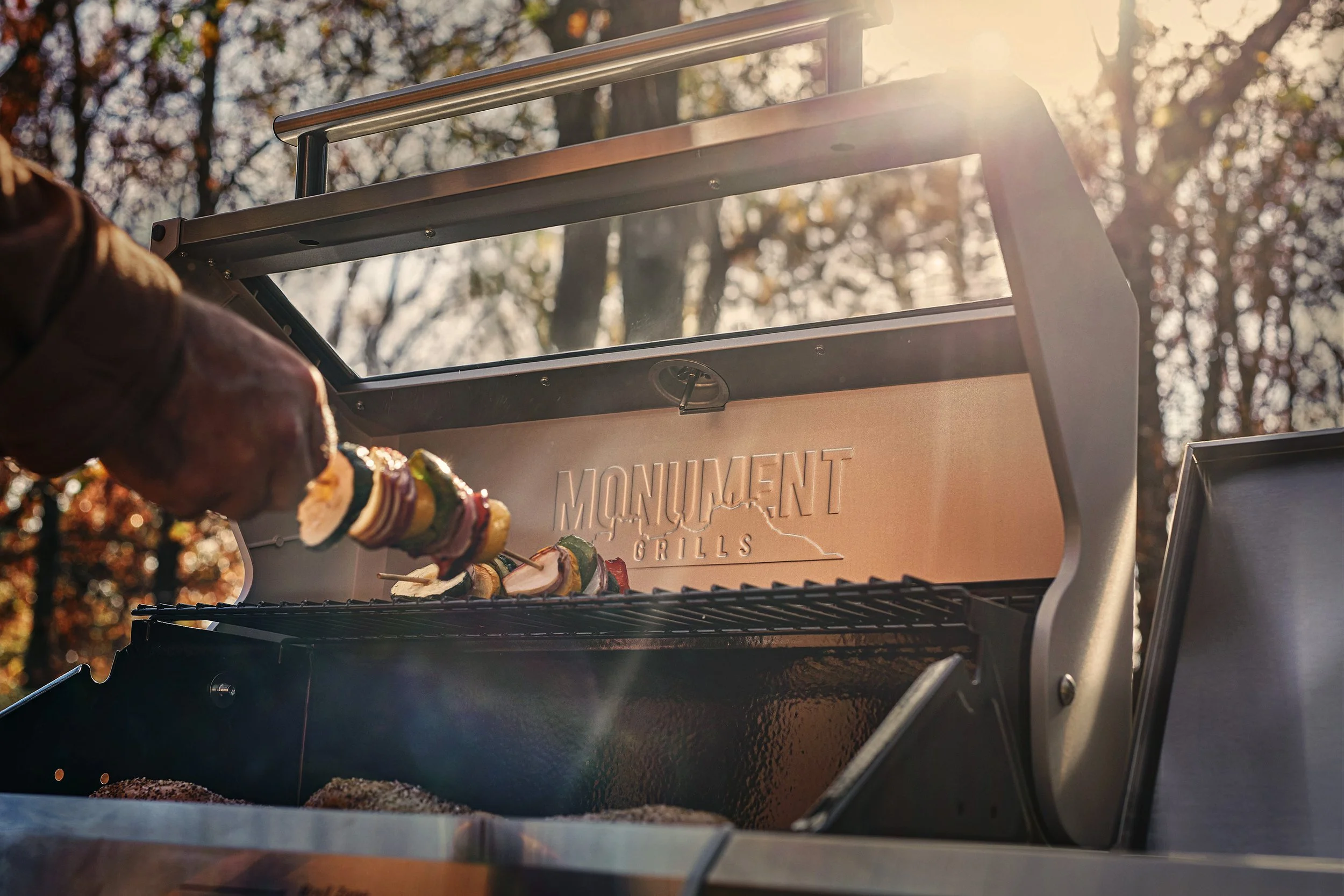 Close-up of a grill with skewers of vegetables being cooked outdoors during sunset.