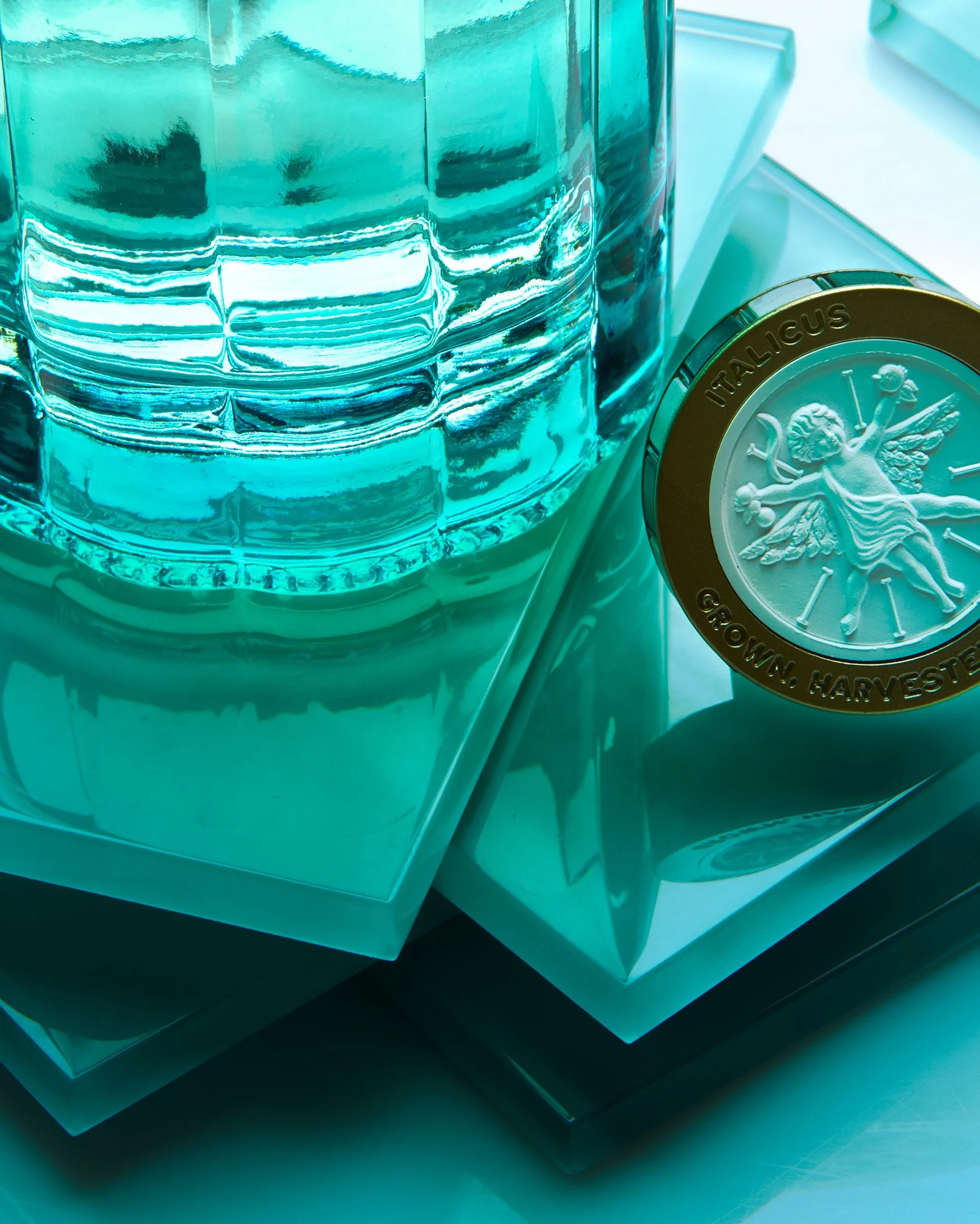 Close-up of a blue glass container, a round coin with a crest and text around it that reads "ITALICUS GROWN. HARVESTED," and a reflective white surface underneath.
