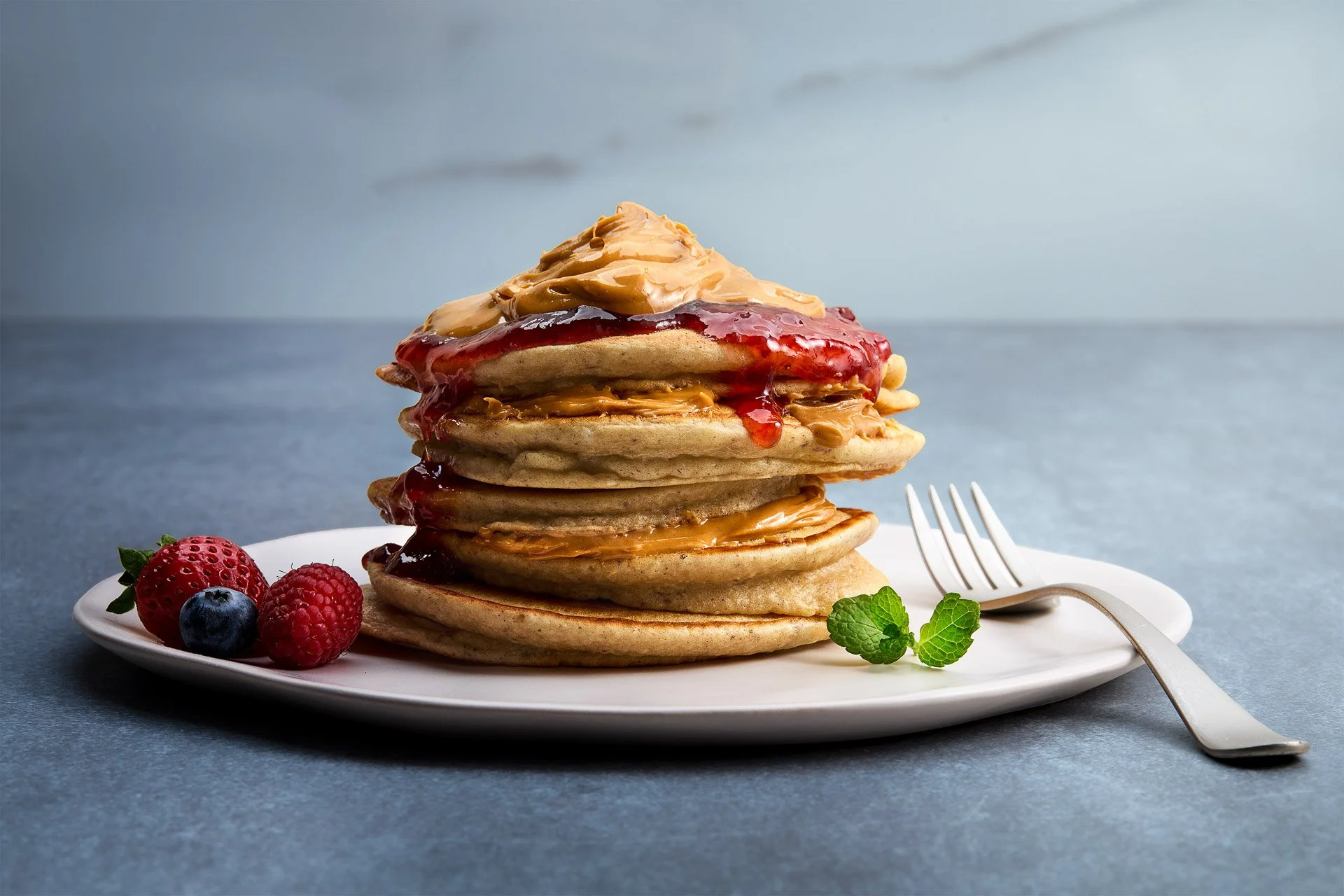 Stack of five pancakes topped with peanut butter, jelly, and syrup on a white plate, garnished with strawberries, raspberries, blueberries, and mint leaves, with a fork on the side, on a blue surface.