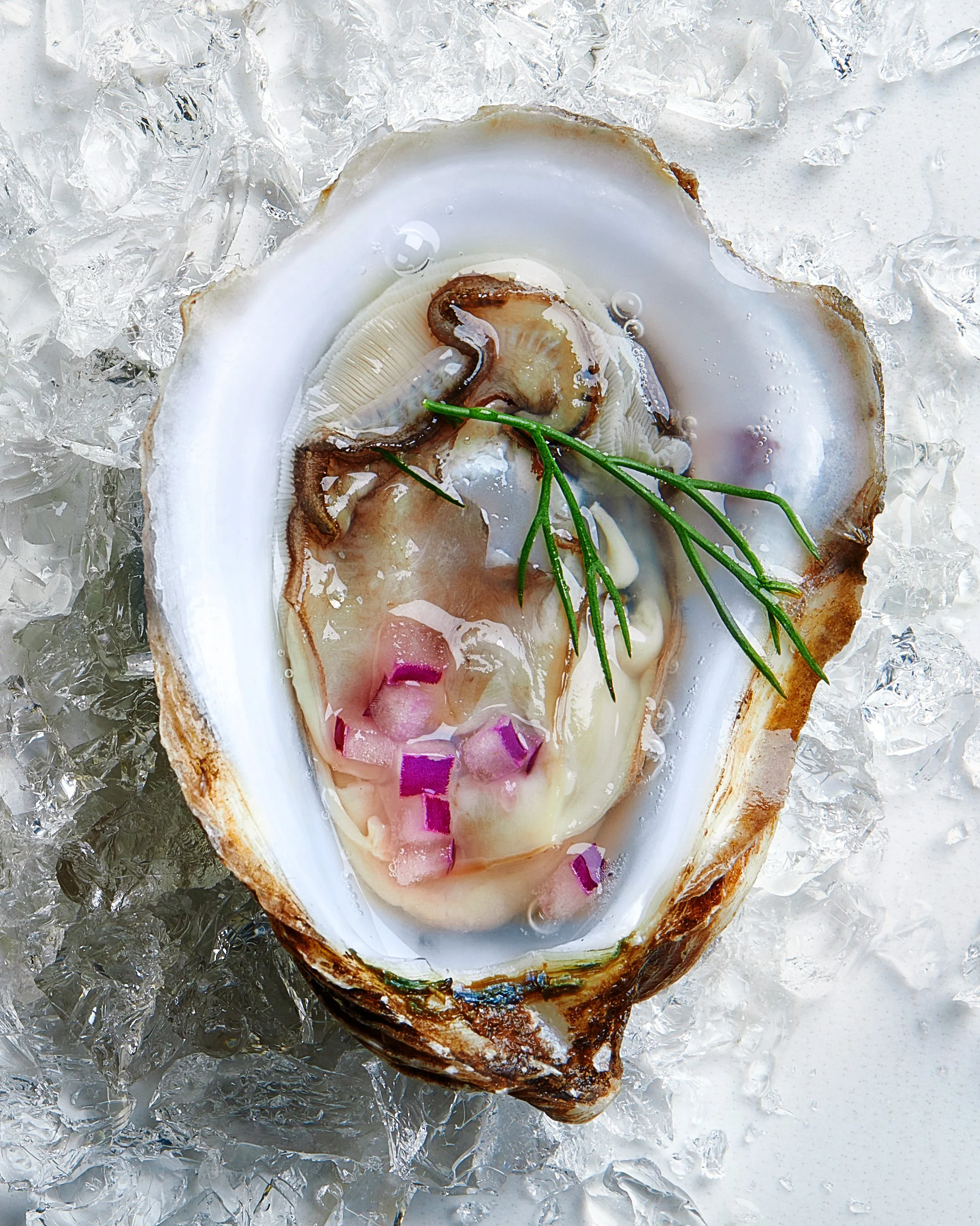 Open oyster shell with raw oyster, garnished with chopped red onions and a sprig of rosemary, on crushed ice.