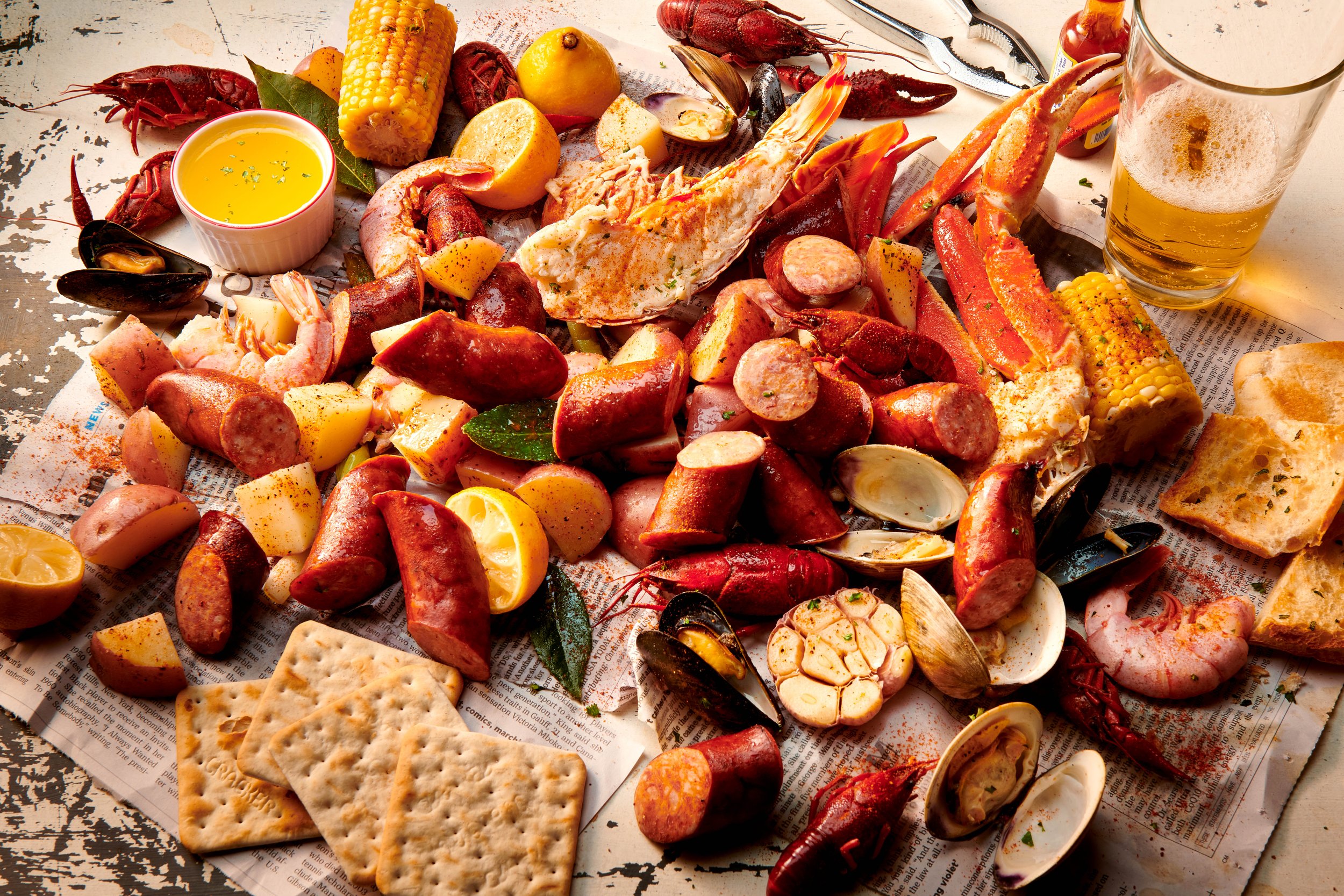 A seafood boil with lobster, crab, shrimp, clams, sausages, corn on the cob, lemon wedges, crackers, and a glass of beer on a paper-lined surface.