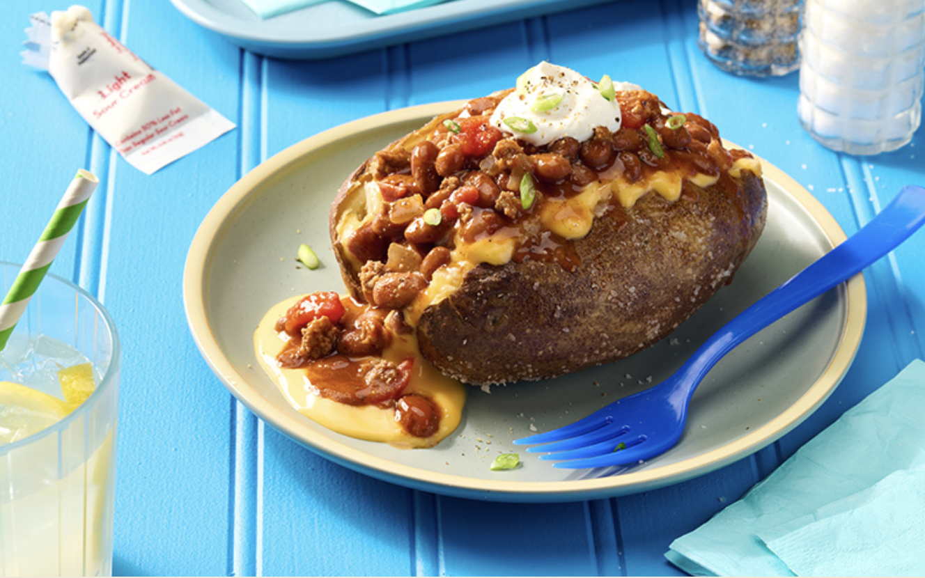 Stuffed baked potato topped with chili, sour cream, and chopped green onions on a green plate, blue fork, lemon drink with straw, salt and pepper shakers, and napkins on a blue table.