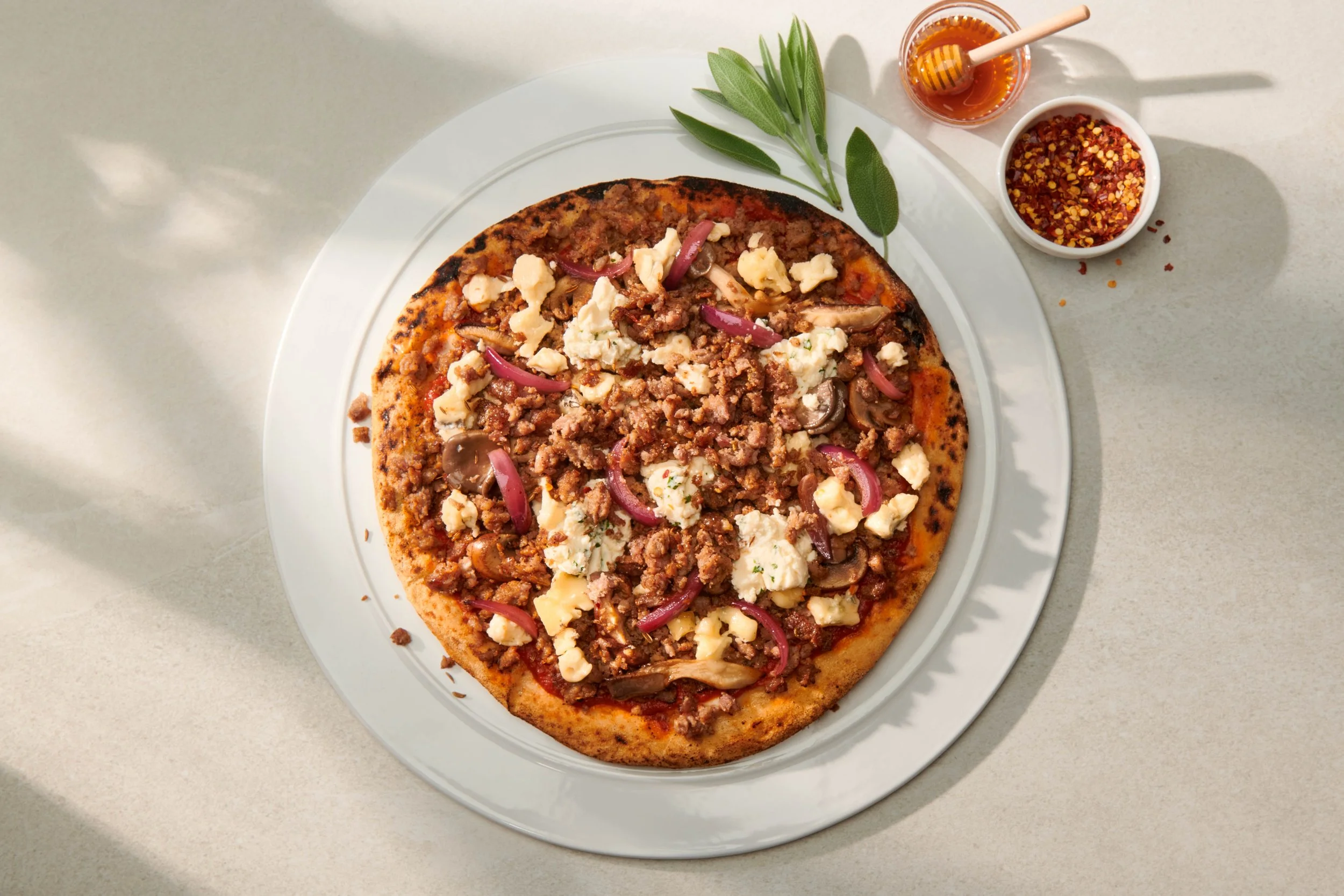 A pizza topped with ground beef, sliced mushrooms, crumbled feta cheese, and pickled red onions on a white plate. Beside the plate, there are a small bowl of red pepper flakes, a small bowl of honey, and a honey dipper. Green sage leaves are placed o