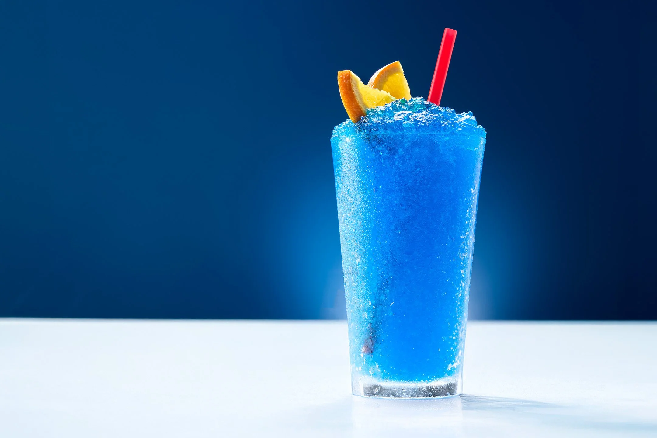 A tall glass of blue frozen tropical drink garnished with orange and lemon wedges and a red straw, on a white surface against a dark blue background.