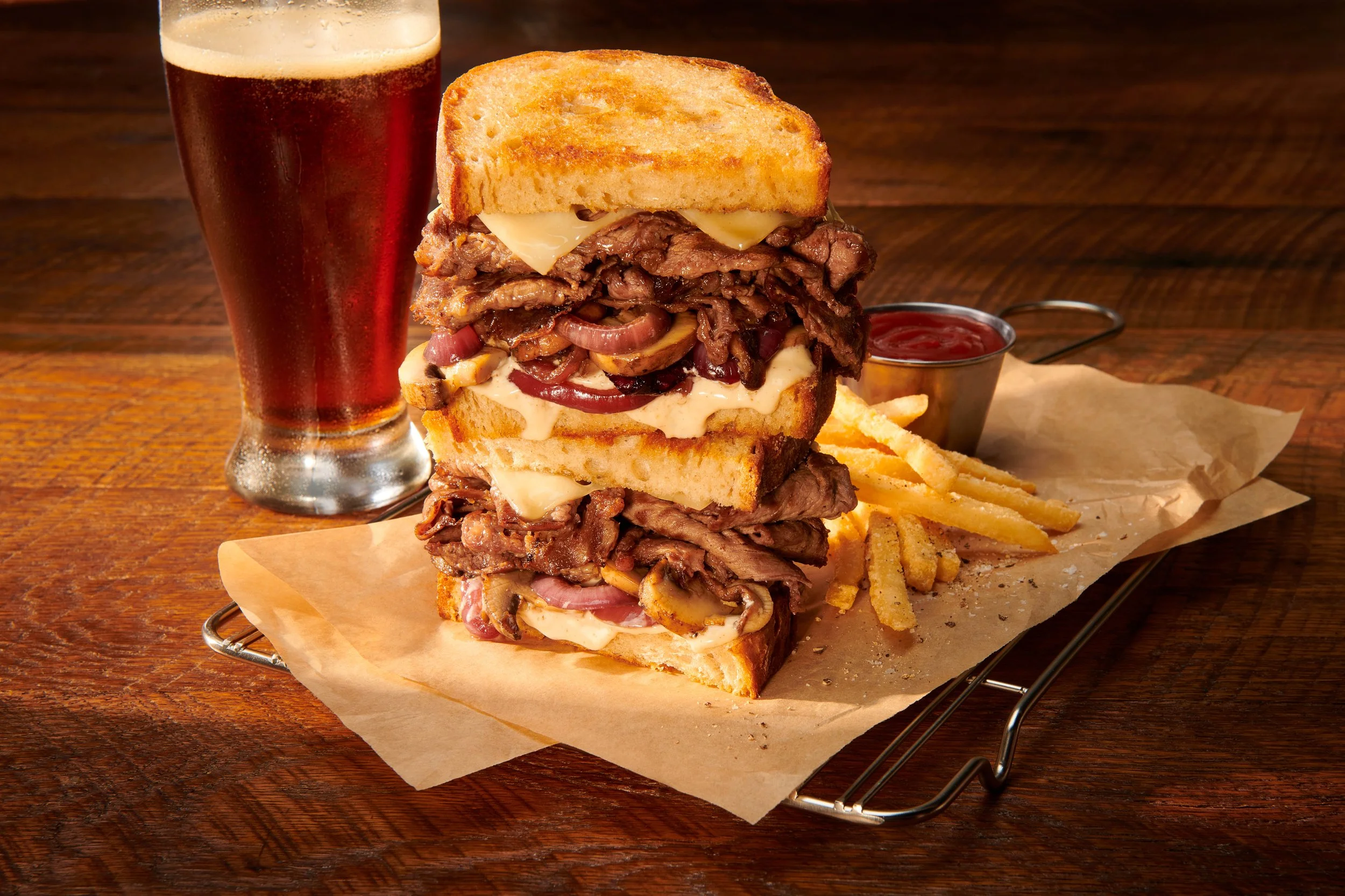 A tall glass of amber beer, a plate with a triple-stacked roast beef and cheese sandwich with fries, ketchup, and dipping sauce on a wooden table.