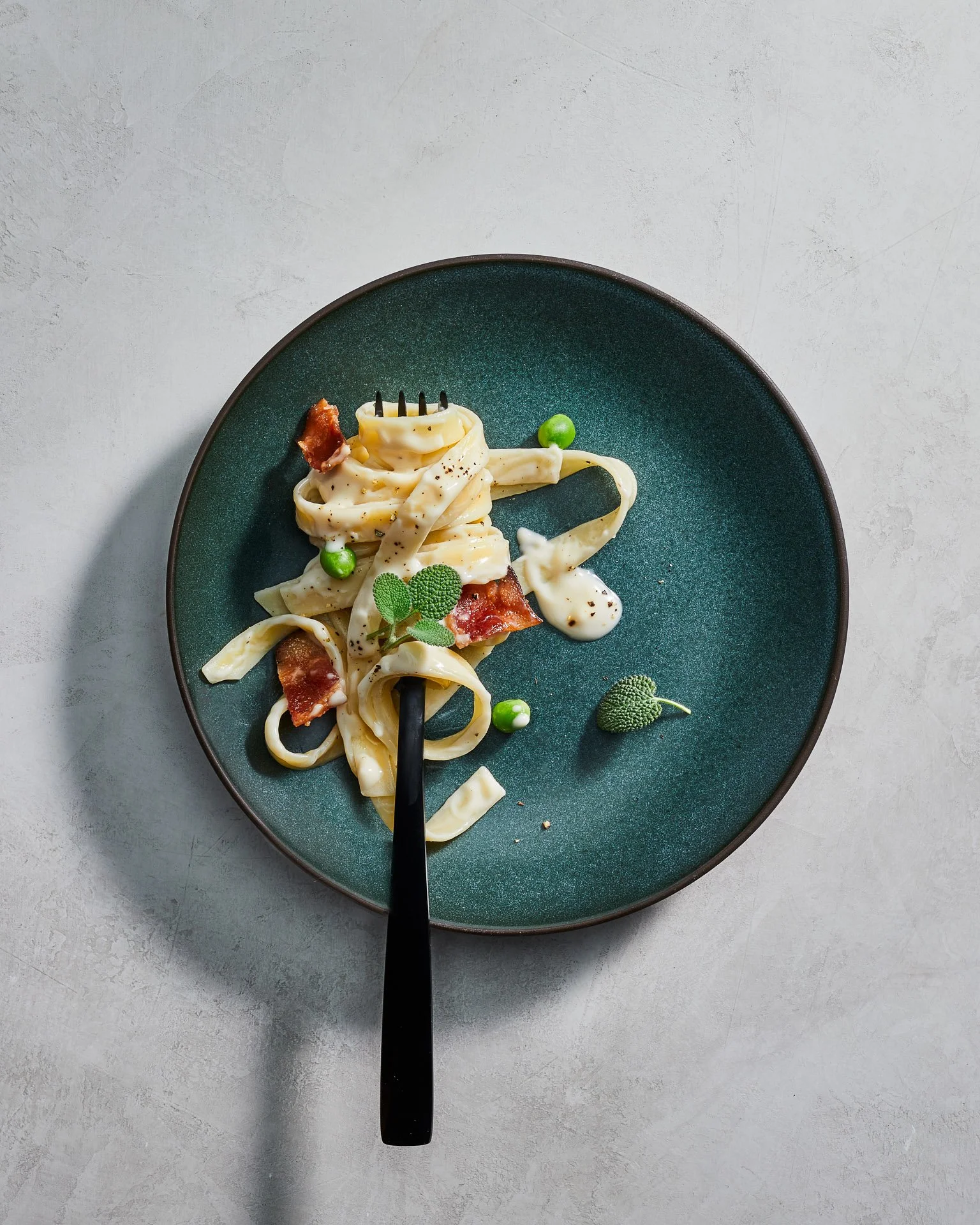 Plate of fettuccine with cream sauce, bacon, peas, and fresh herbs, presented with a black fork on a dark green plate.