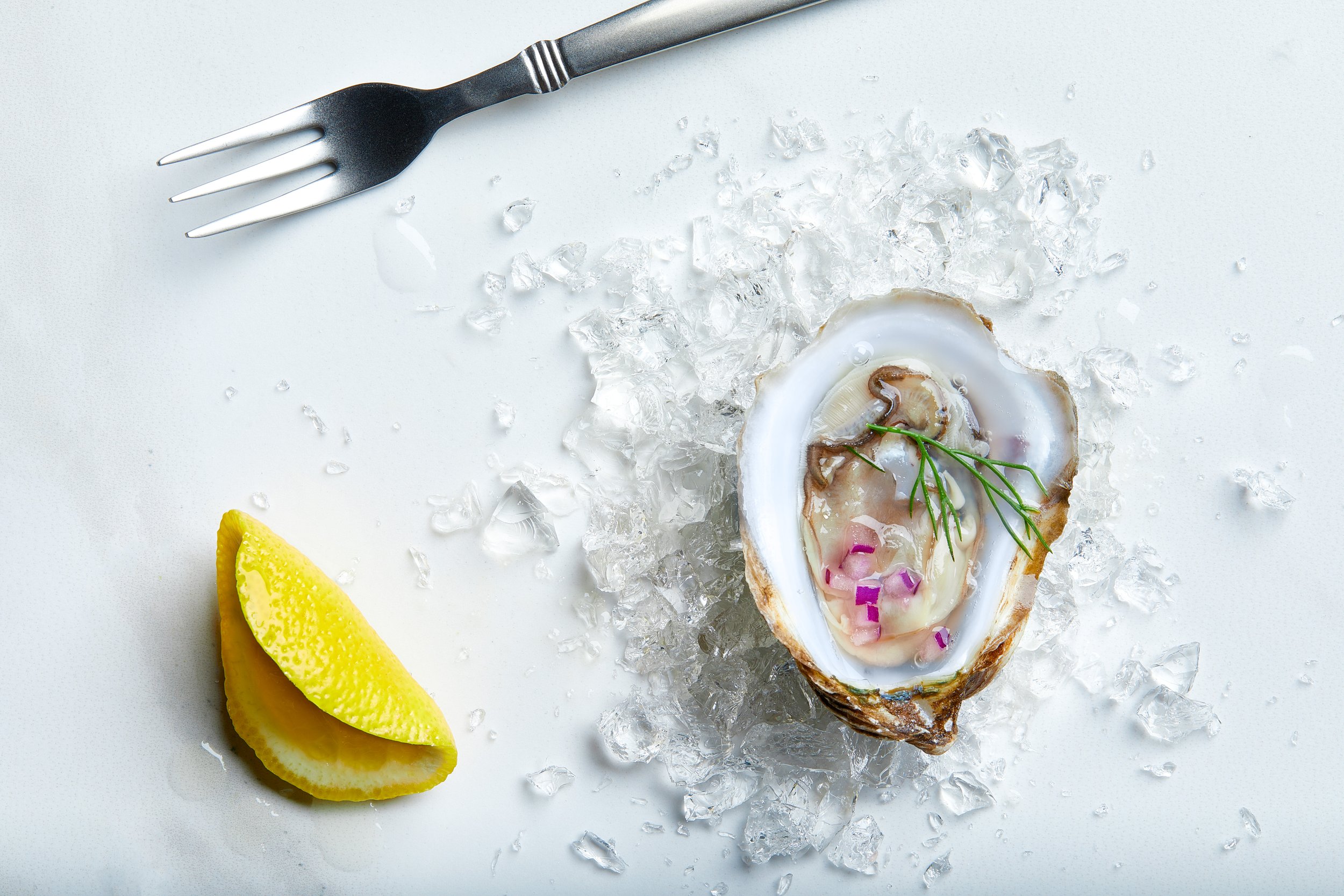 Raw oyster on a half shell, garnished with herbs, surrounded by crushed ice, with a lemon wedge and a fork nearby.