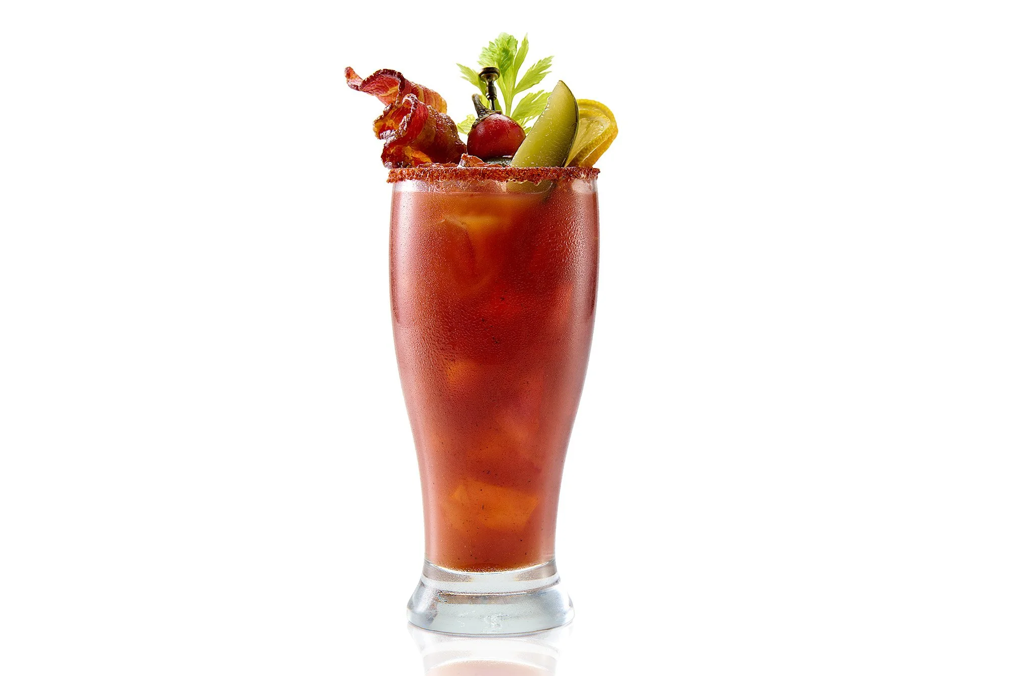 A tall glass of Bloody Mary cocktail garnished with bacon strip, cherry tomato, lime wedge, celery root, and leafy greens on a white background.