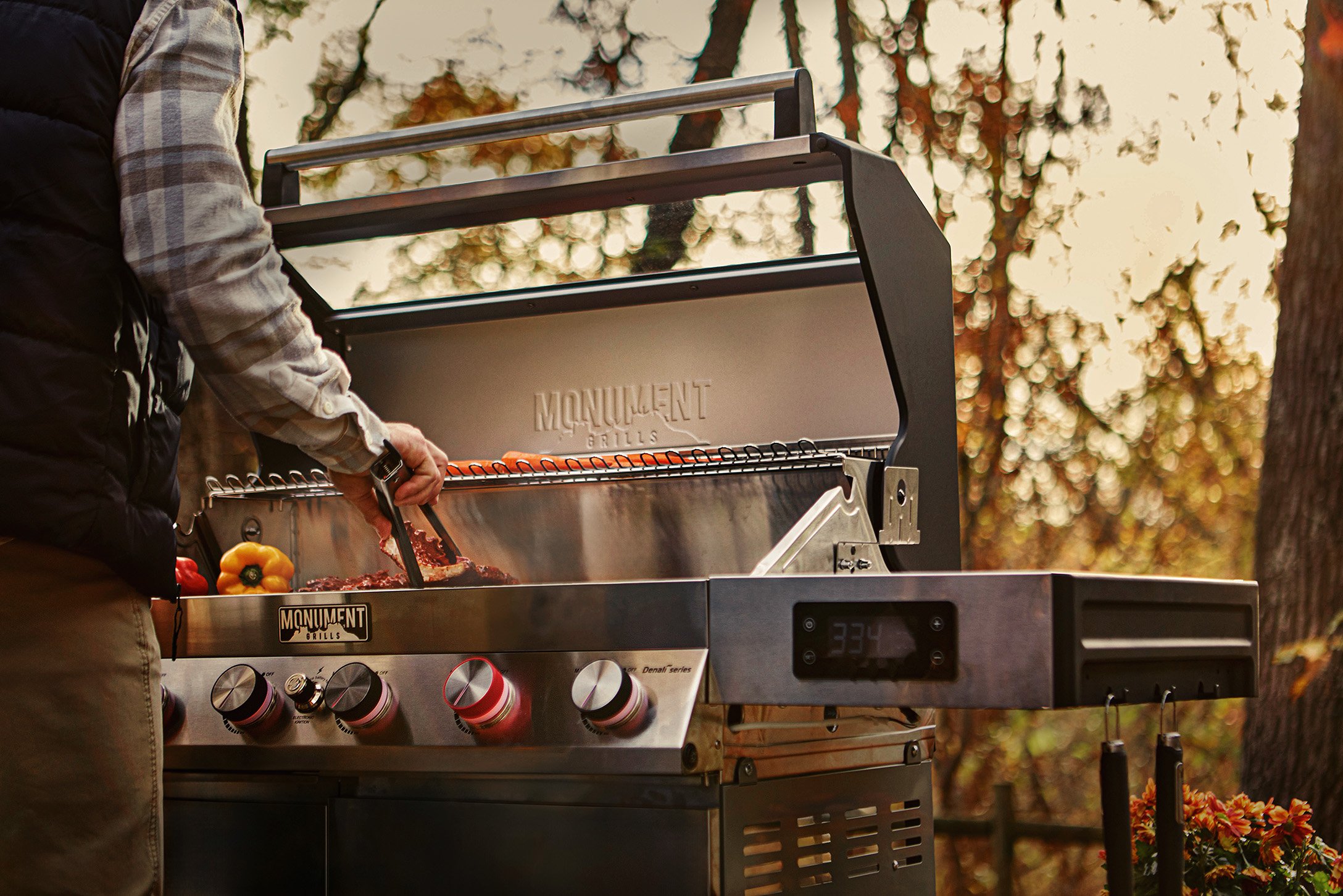 Person grilling hot dogs and vegetables on a barbecue grill outdoors during sunset.