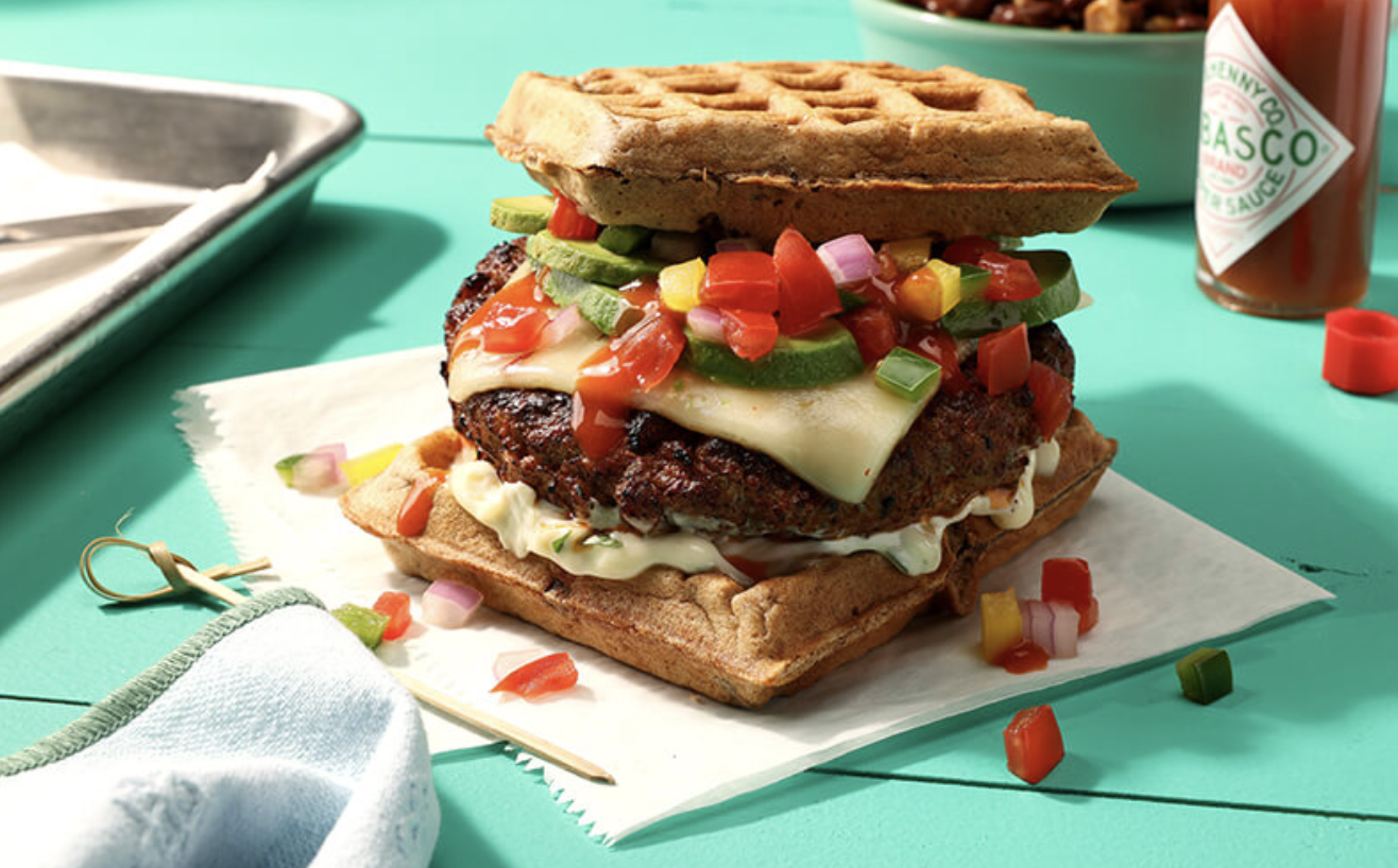 A large homemade burger with a waffle bun, topped with cheese, vegetables, and ketchup, on a piece of paper on a teal surface.