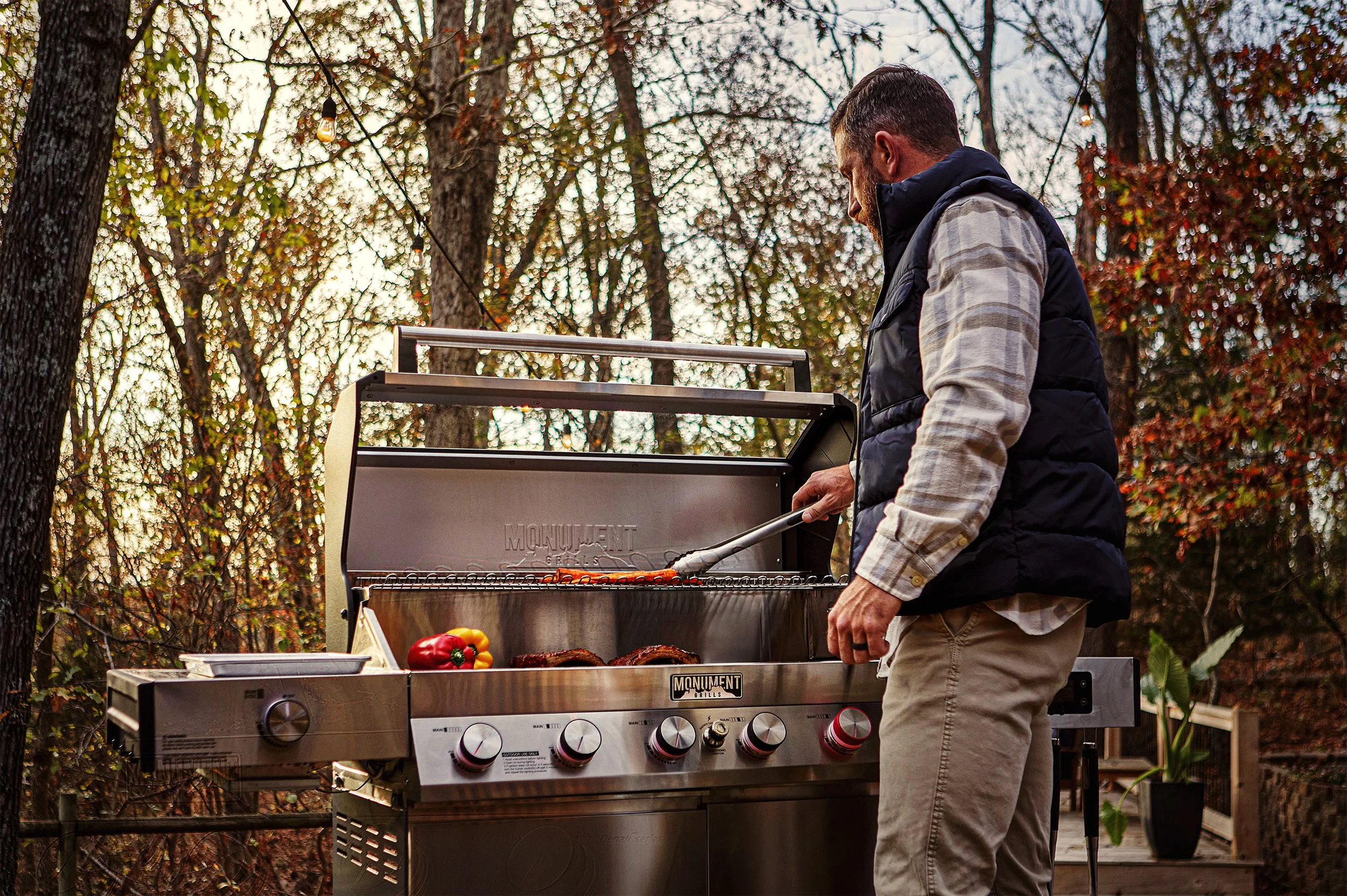 A man grilling vegetables and burgers outdoors on a stainless steel grill surrounded by trees with autumn leaves.