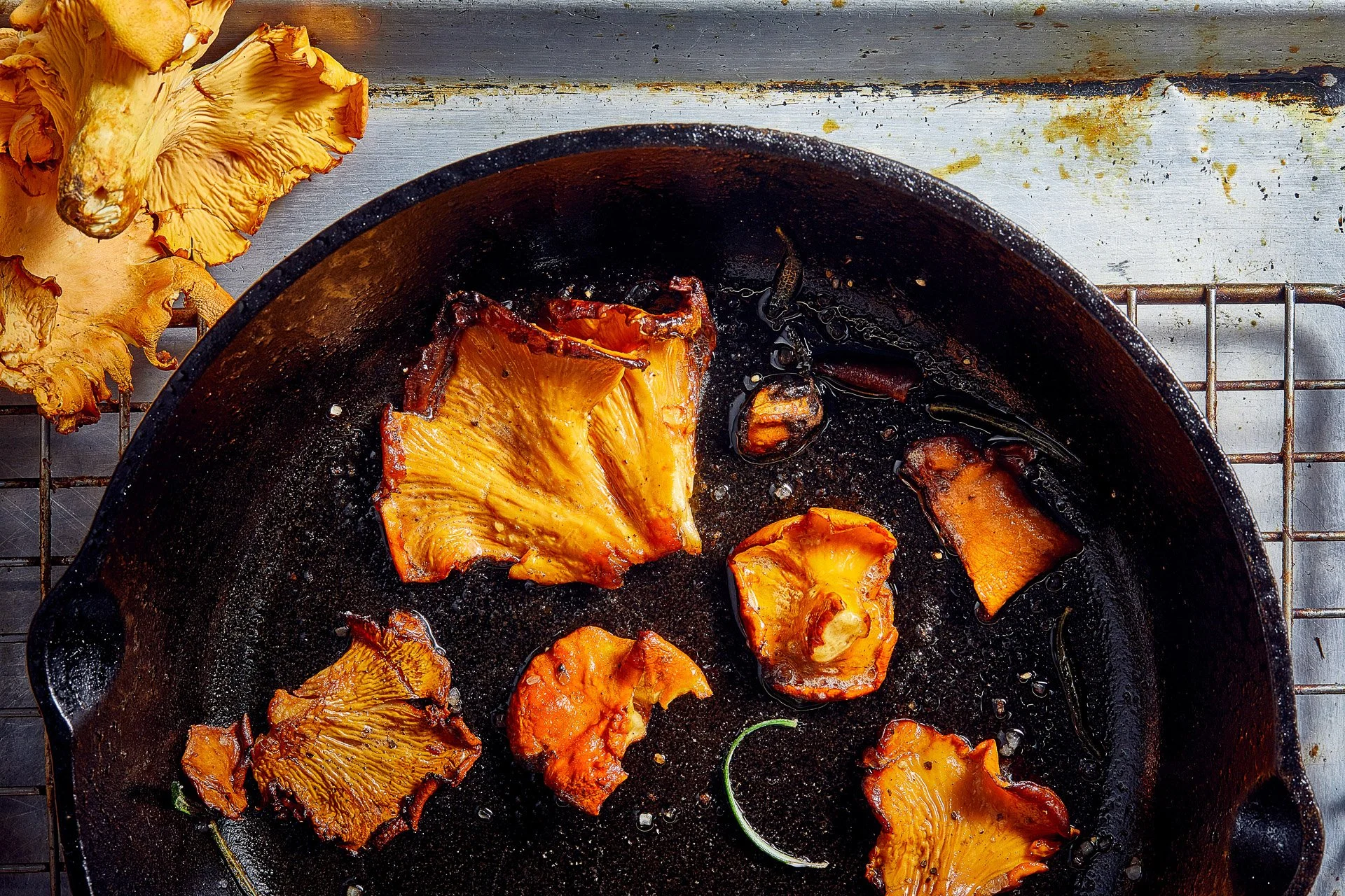 Chanterelle mushrooms cooking in a cast iron skillet, with a few raw mushrooms nearby on a wire rack.