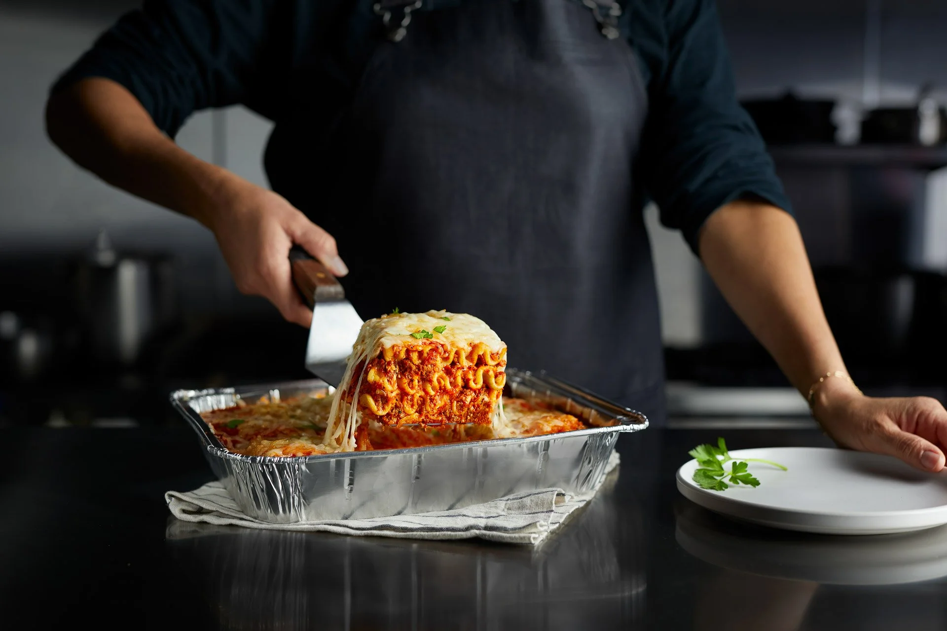 Person lifting a slice of lasagna from a foil tray with a spatula, holding a plate with parsley garnish nearby.