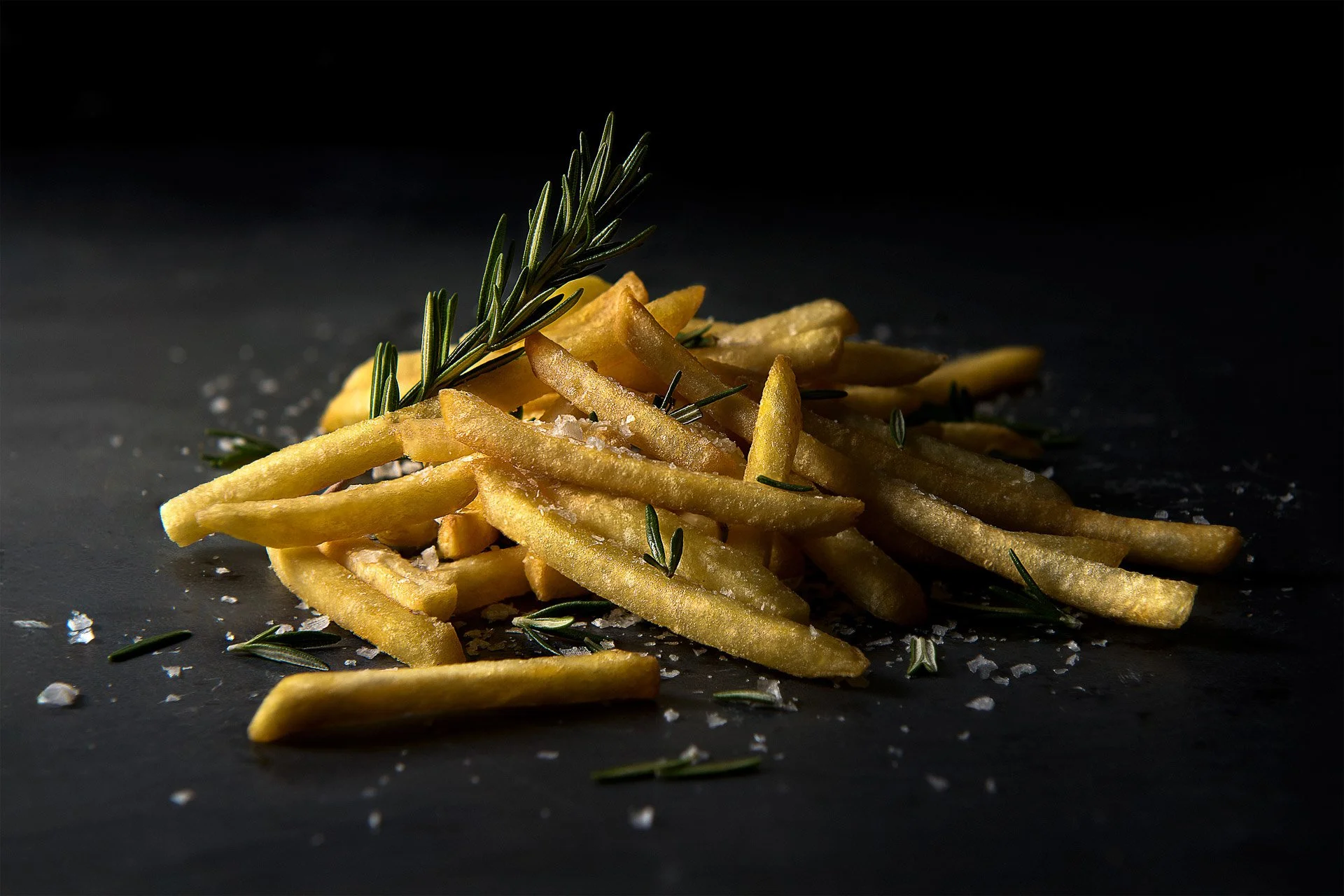 French fries garnished with fresh rosemary and sprinkled with coarse salt on a dark surface.
