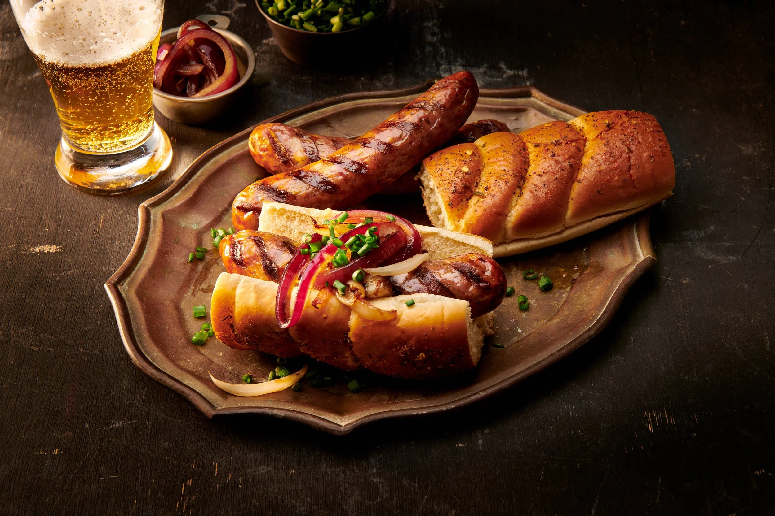 Traditional German sausage platter with grilled sausages, sliced bread, and onion and chive garnishes, served with a glass of beer.