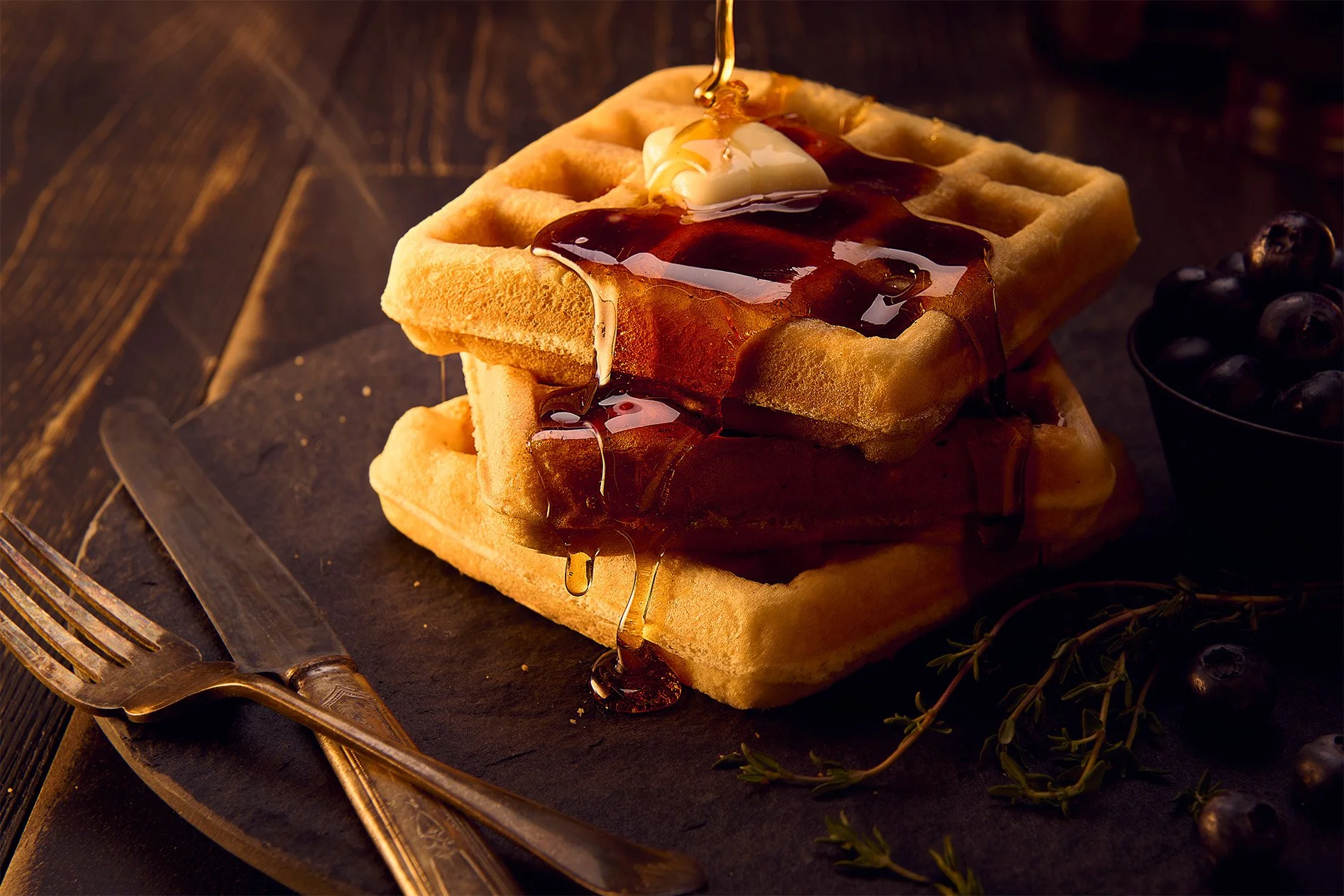 Two golden Belgian waffles stacked with syrup and a dollop of butter, served on a dark slate with blueberries, a fork, and knife nearby.