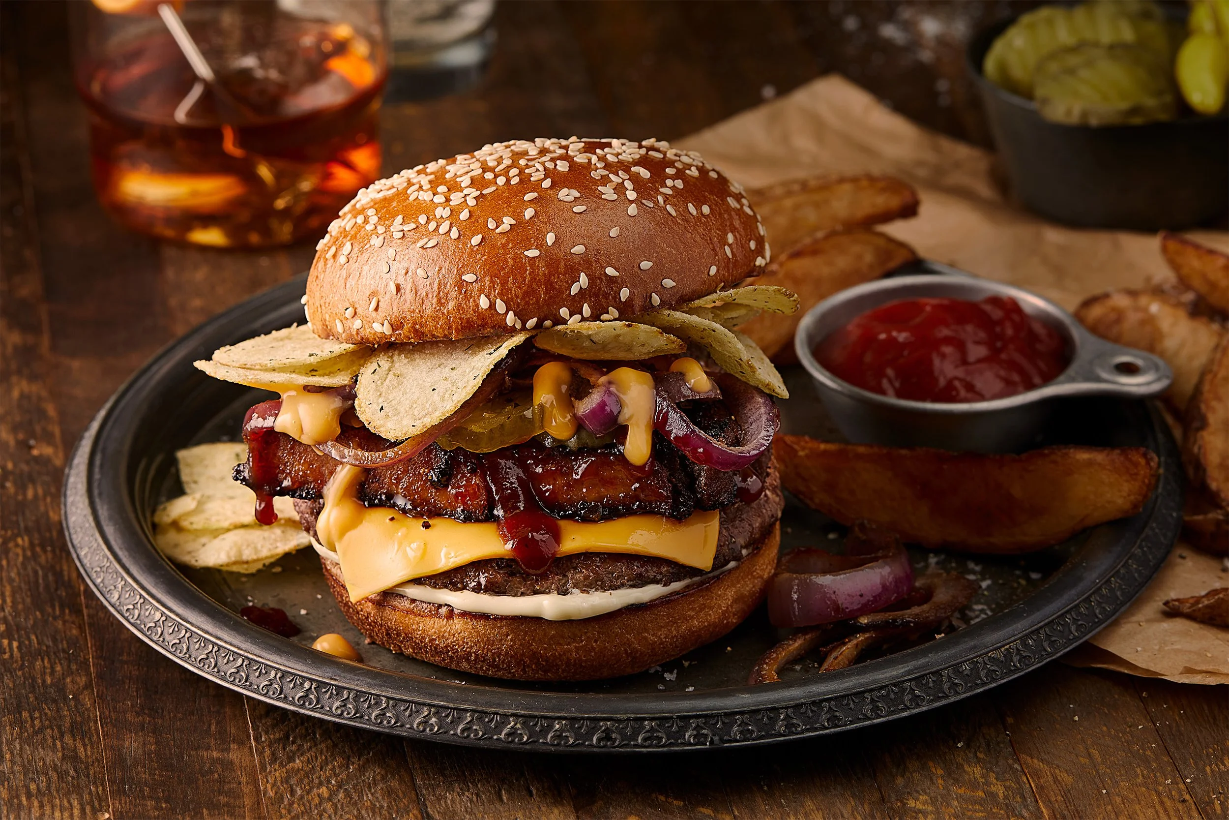 A gourmet burger with cheese, bacon, pickles, onions, and condiments in a sesame seed bun, served with potato wedges and a side of ketchup on a black plate.