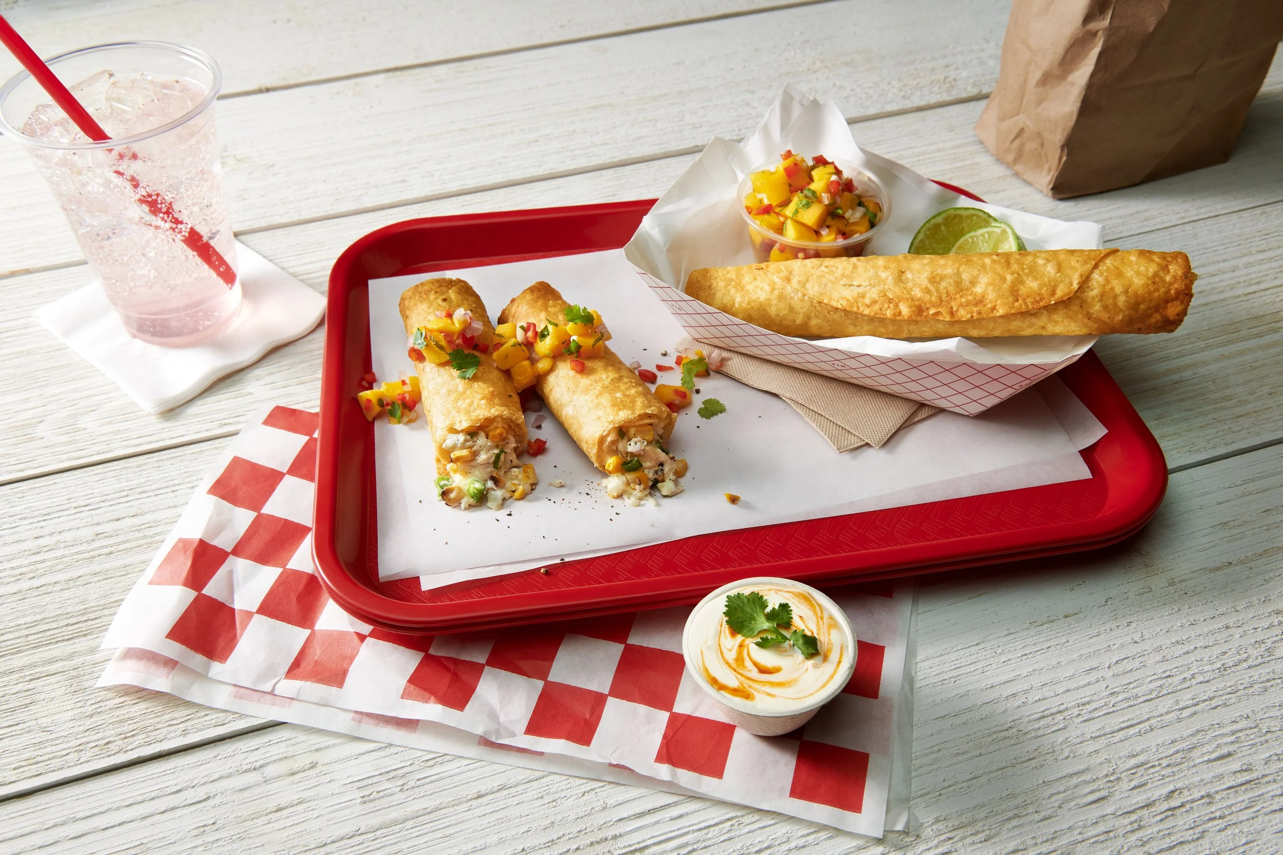 Tray of Mexican food including a large taquito, two smaller taquitos with toppings, a fried taquito in a basket, lime wedge, small bowl of mango salsa, and a container of sour cream on a wooden table.