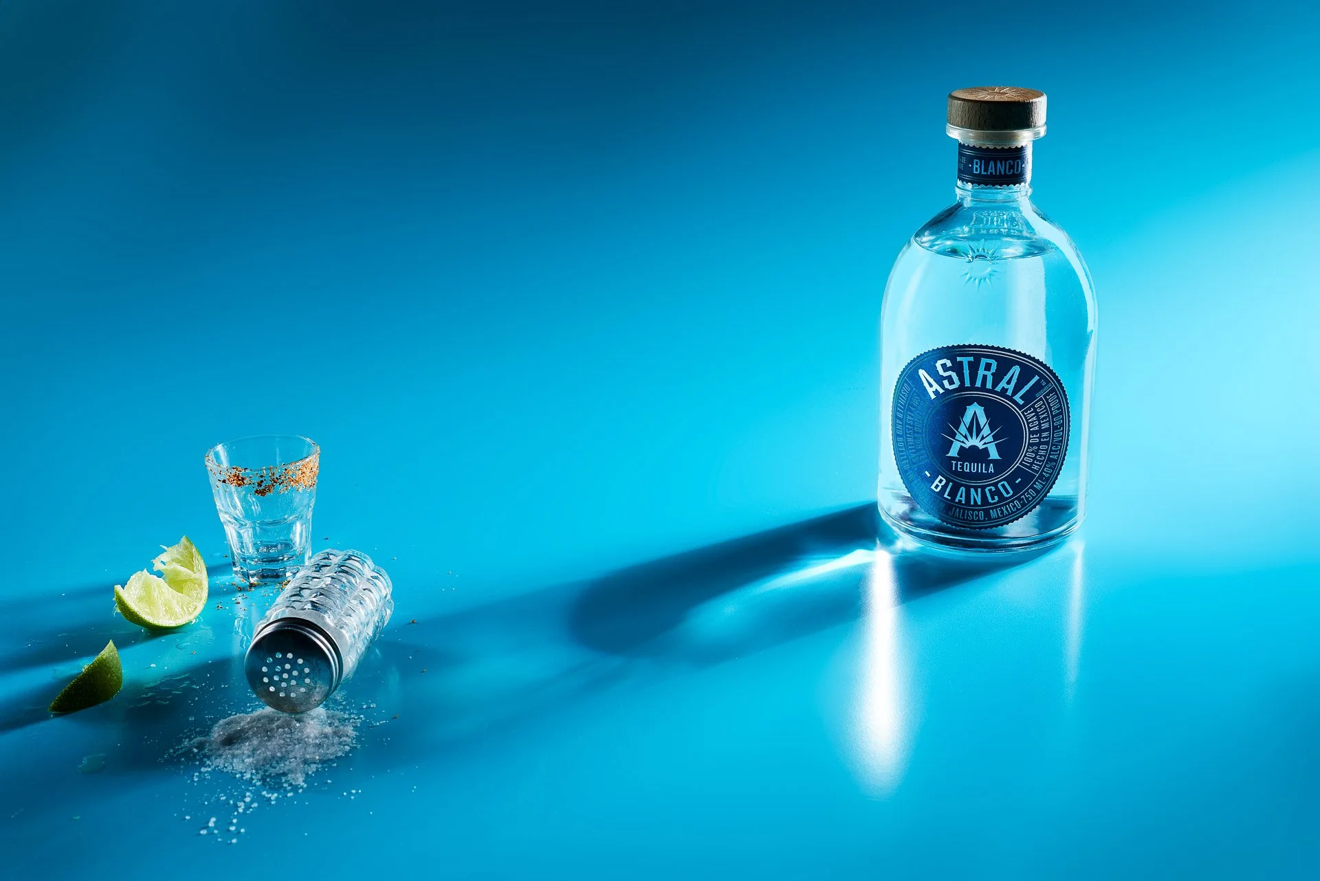 Bottle of Astral Blanco Tequila on a blue surface with a shot glass rimmed with spices, lime wedges, and a spilled salt shaker.