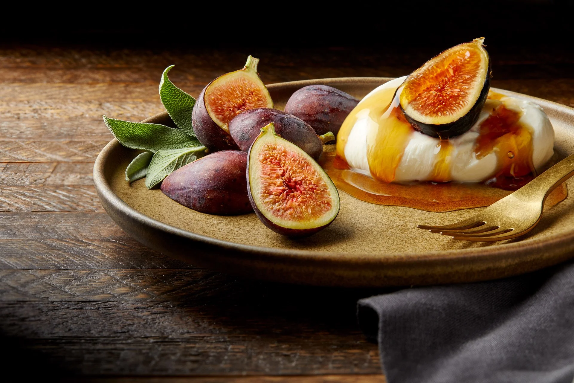 Fresh figs and creamy cheese drizzled with honey on a ceramic plate with a gold fork, garnished with sage leaves, set on a wooden table with a dark fabric napkin.