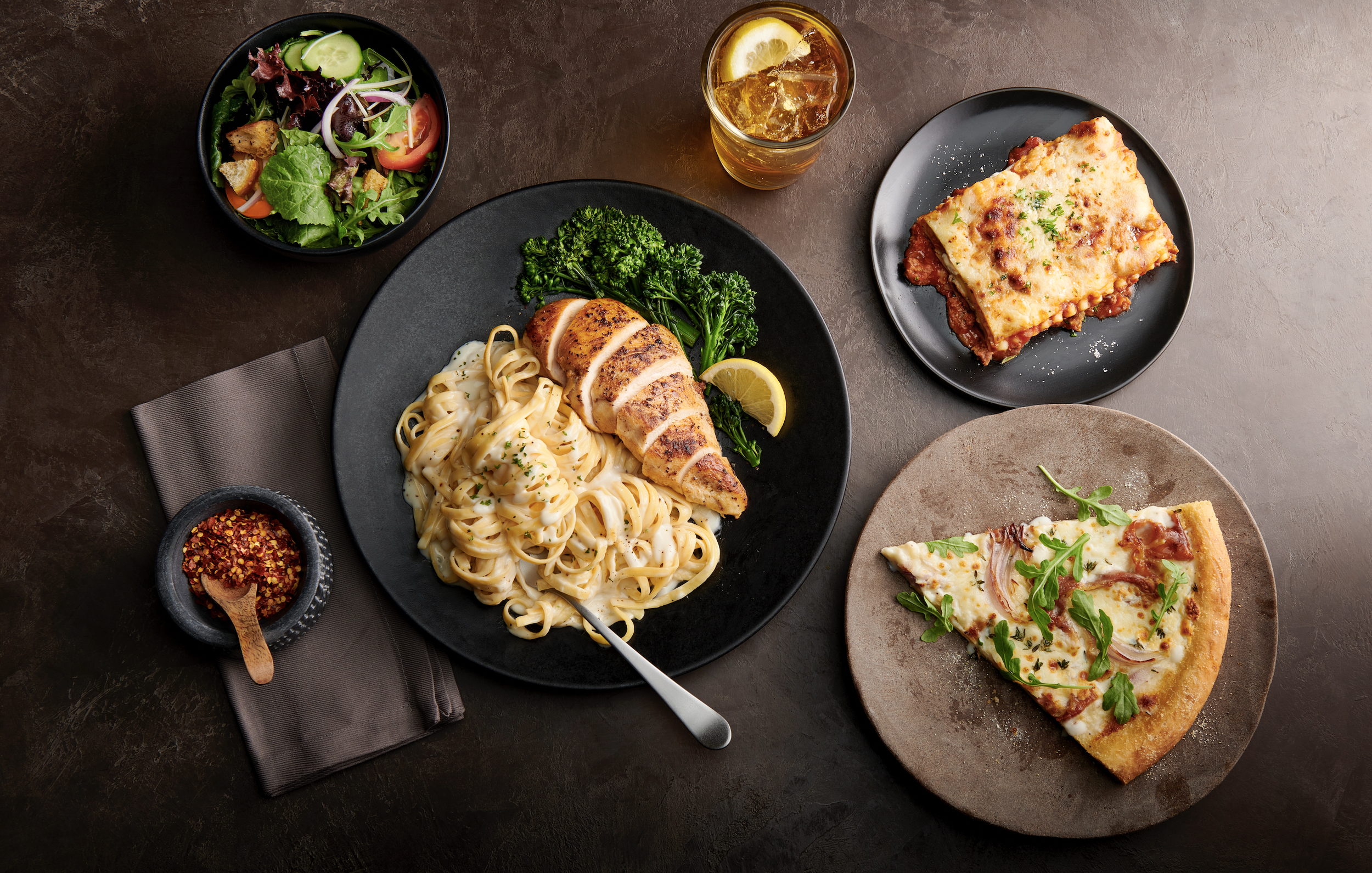A top-down view of a dinner spread with a bowl of salad, a grilled chicken Alfredo pasta dish, a glass of iced tea, a slice of pizza, and a serving of lasagna on separate black and ceramic plates, all placed on a dark surface.