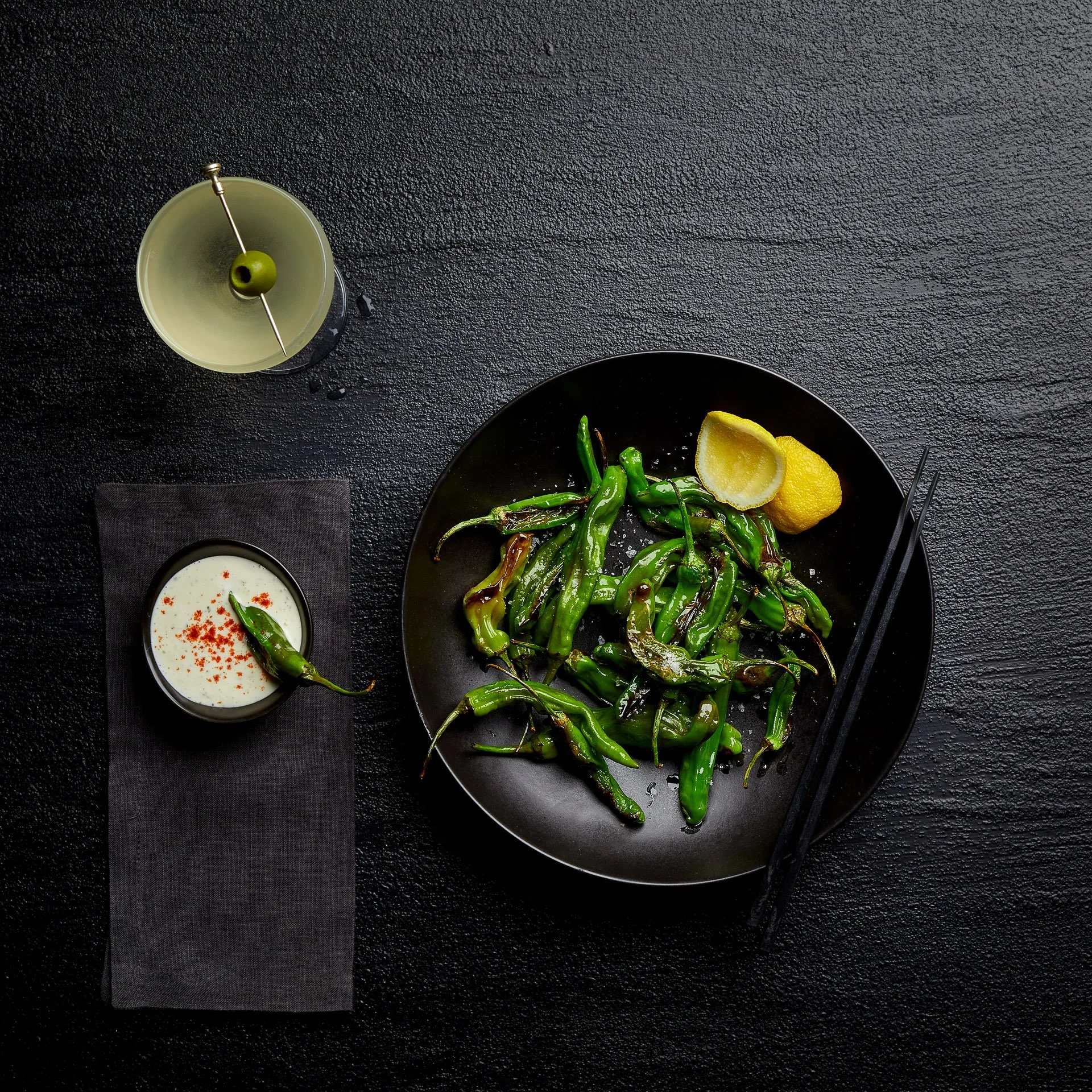 Plate of roasted shishito peppers with lemon wedges, a drink with an olive, black chopsticks, and a bowl of dipping sauce on a dark surface.