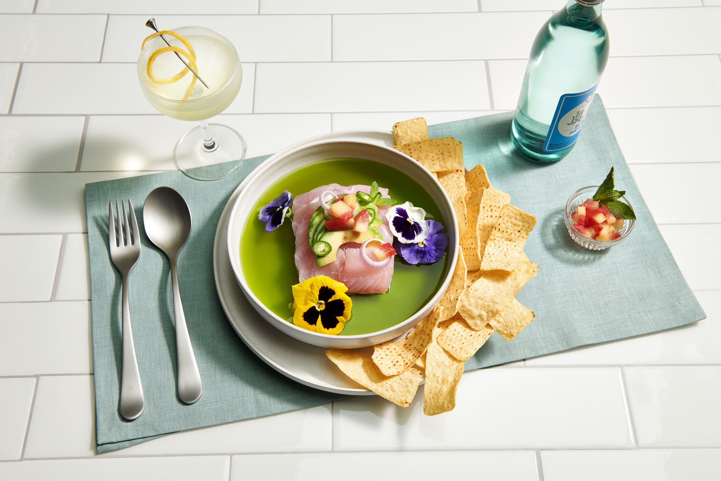 A plate of ceviche garnished with edible flowers, served with tortilla chips. There is a small bowl of fruit salsa, a bottle of sparkling water, and a cocktail with lemon twist on a white tiled surface with a teal placemat.